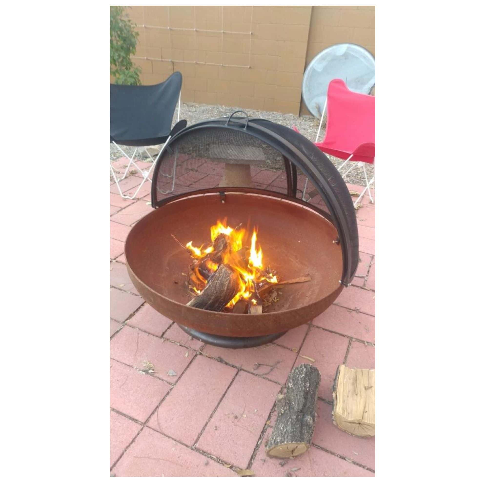 A round metal fire pit with a black mesh screen and lid, containing a fire with bright orange flames, set on a red brick patio outdoors.