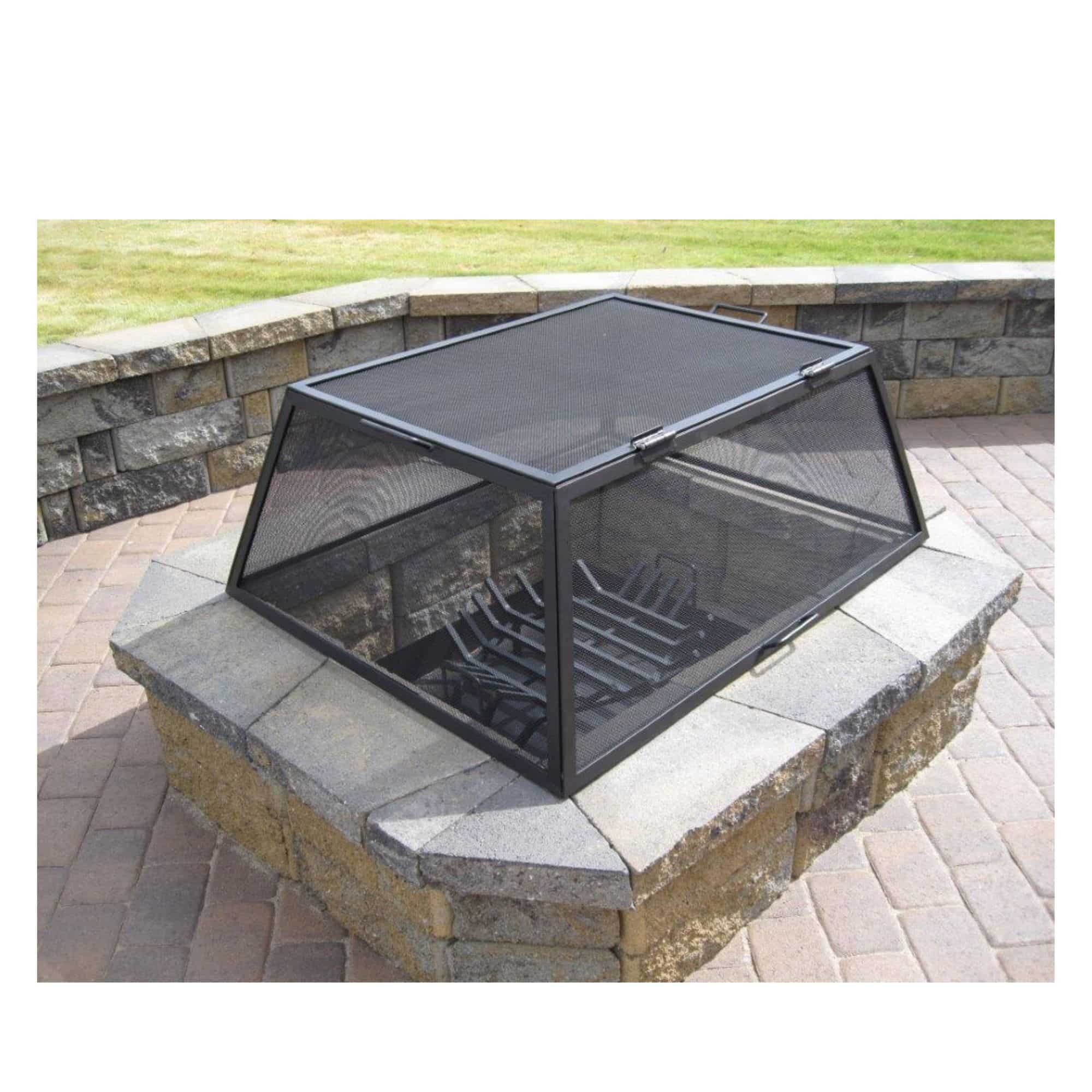Black metal fire pit cover with mesh screen and interior fire grate, shown placed on a stone fire pit outdoors on a paved patio with grass in the background.
