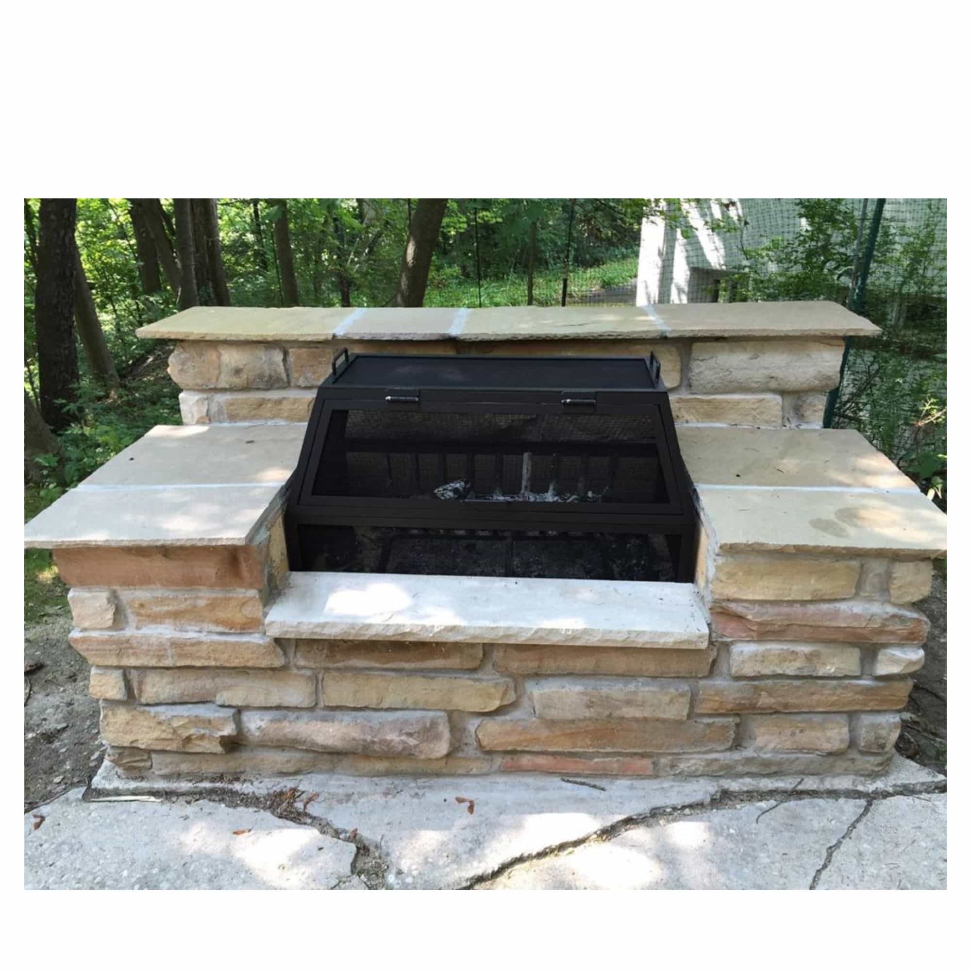 A black fire pit screen set into a stone outdoor fireplace with tan and beige natural stones, photographed outdoors with trees and a concrete ground visible.