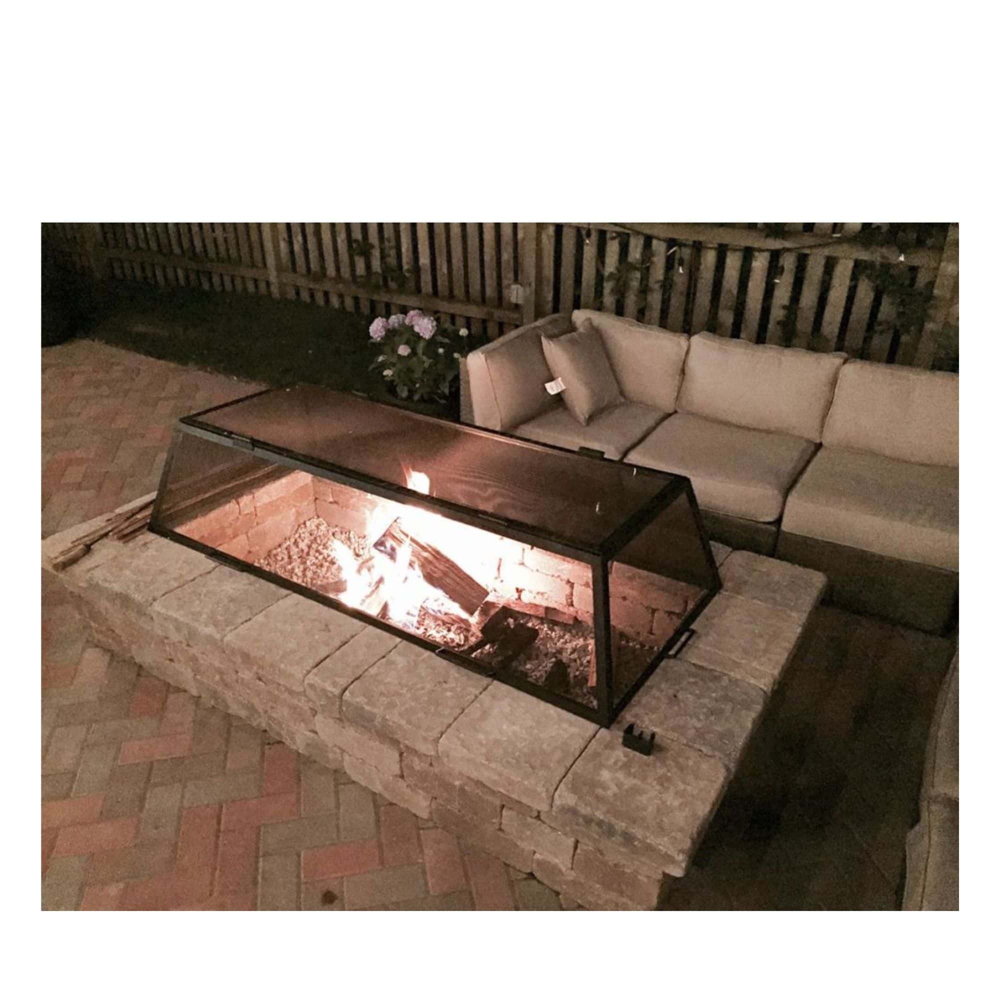 A rectangular outdoor fire pit screen with a black metal frame, shown lit with visible flames on a stone patio next to a beige outdoor sofa at night.