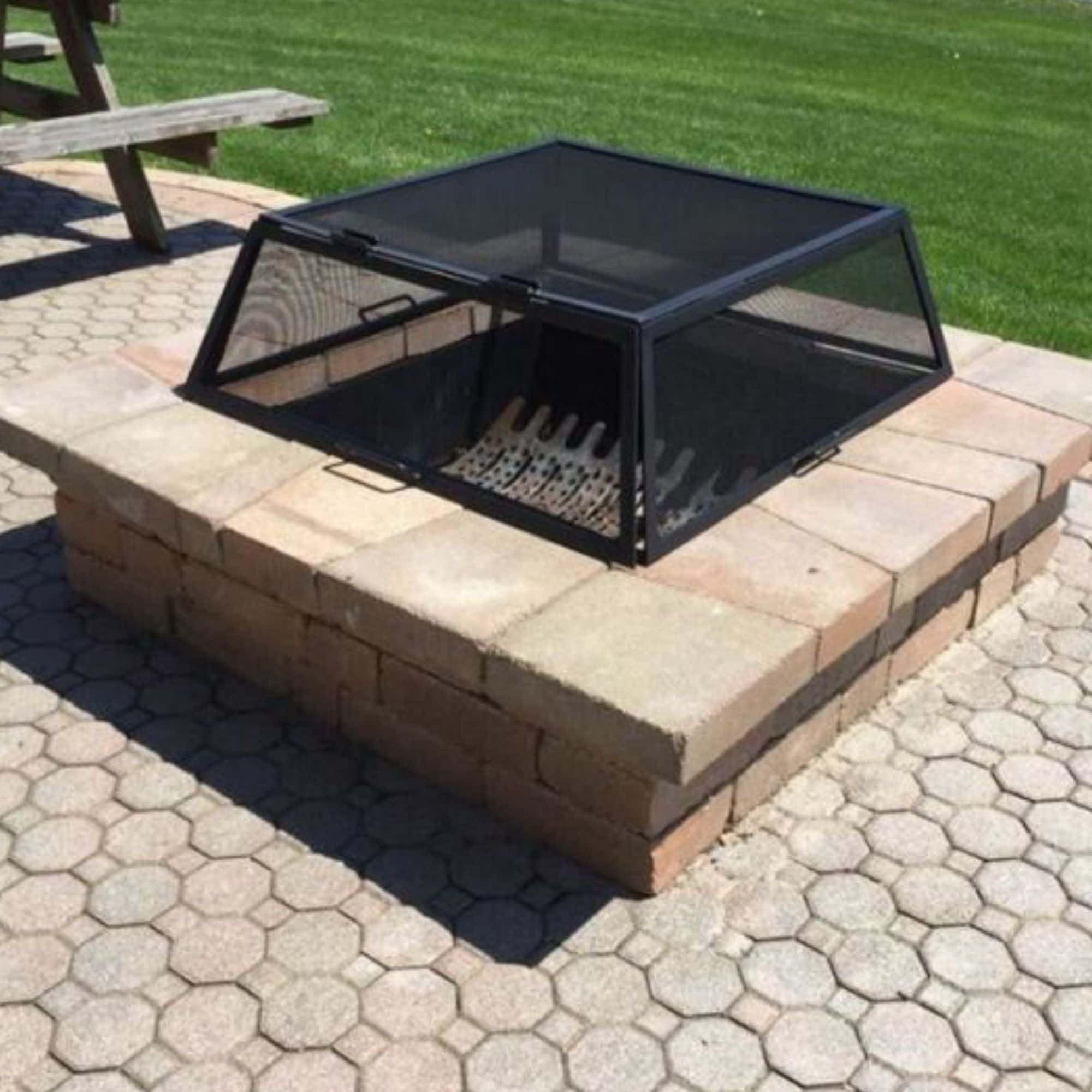 A black metal mesh fire pit cover with a hinged lid, shown installed on top of a brick fire pit outdoors on a paved patio with grass in the background.