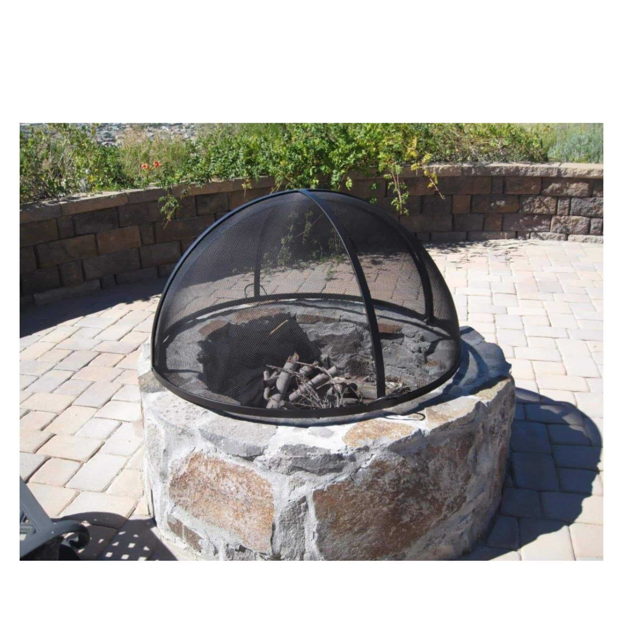 A stone fire pit with a black mesh dome screen cover, set on a paved patio with a stone wall and greenery in the background.