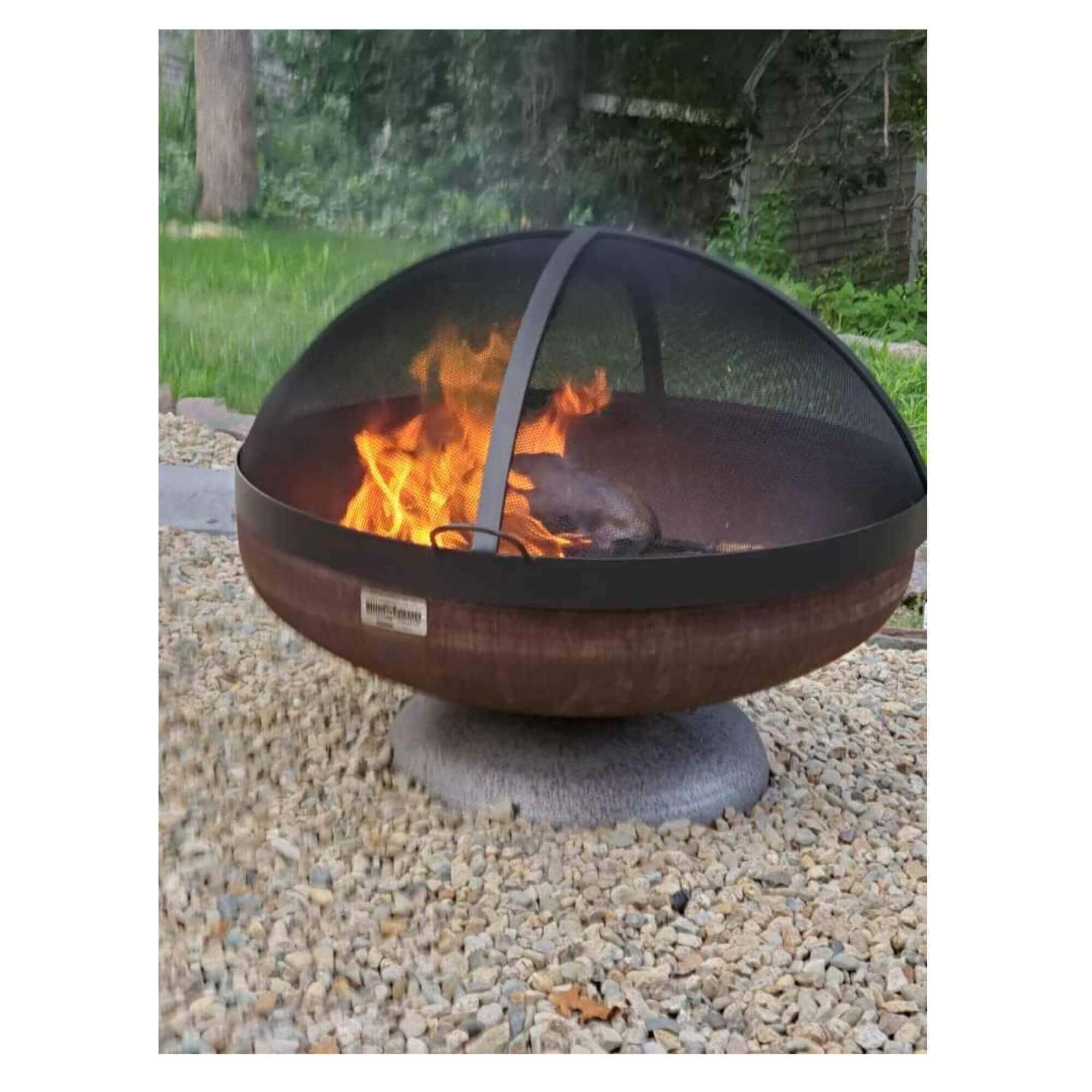 A round metal fire pit with a dark mesh dome cover and visible orange flames, sitting on a stone base over gravel outdoors.