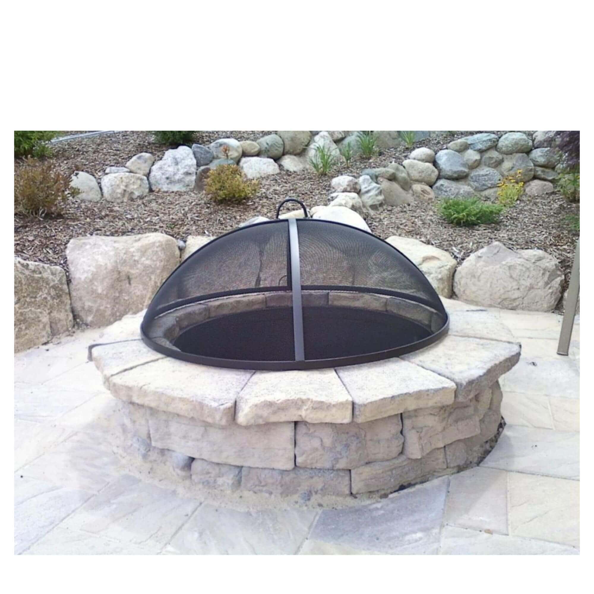 A round stone fire pit with a black mesh dome cover, set on a paved patio in an outdoor backyard landscape with rocks and mulch.