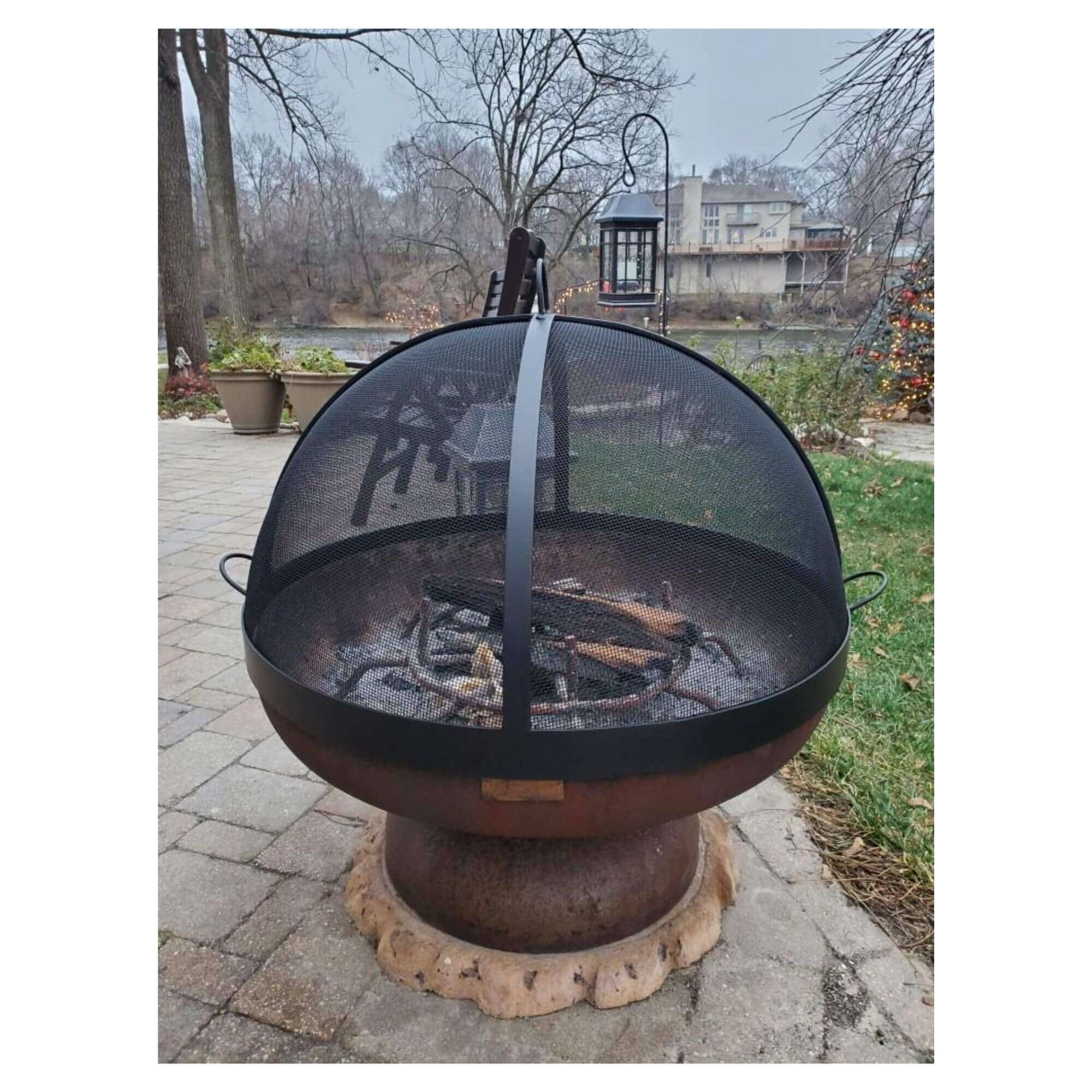 A round outdoor fire pit with a black mesh dome cover and a rusted metal base, shown on a stone patio in a backyard setting with trees and a house in the background.