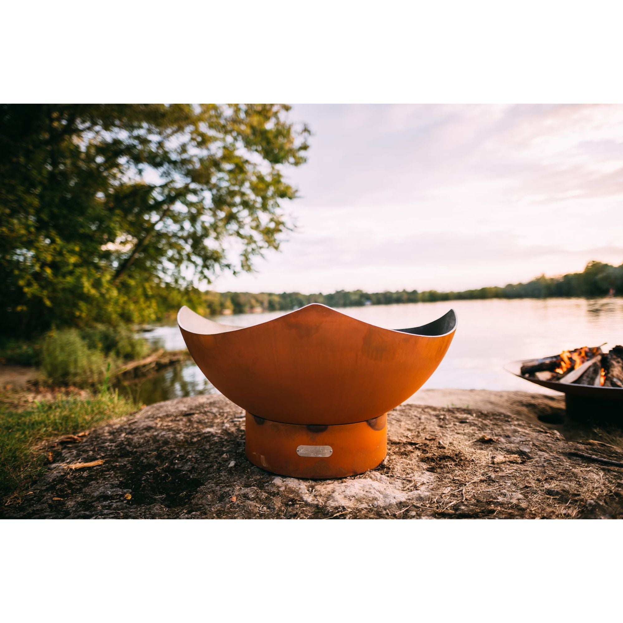 Orange fire pit on a rock by a lake with trees in the background