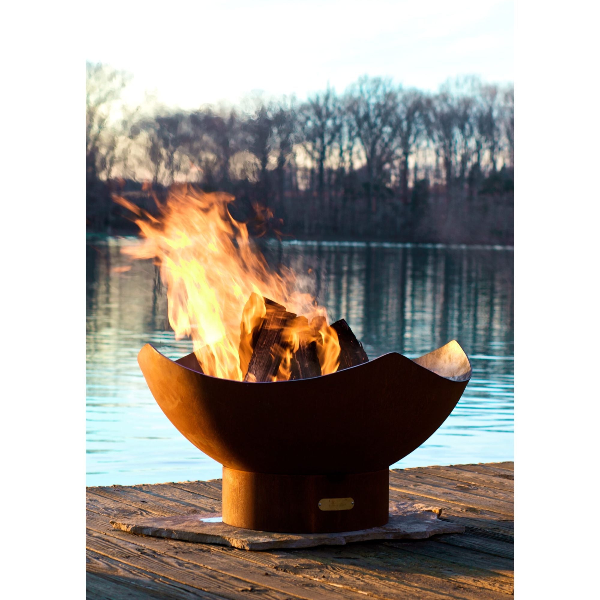 Fire pit on a wooden dock by a lake with flames burning brightly.