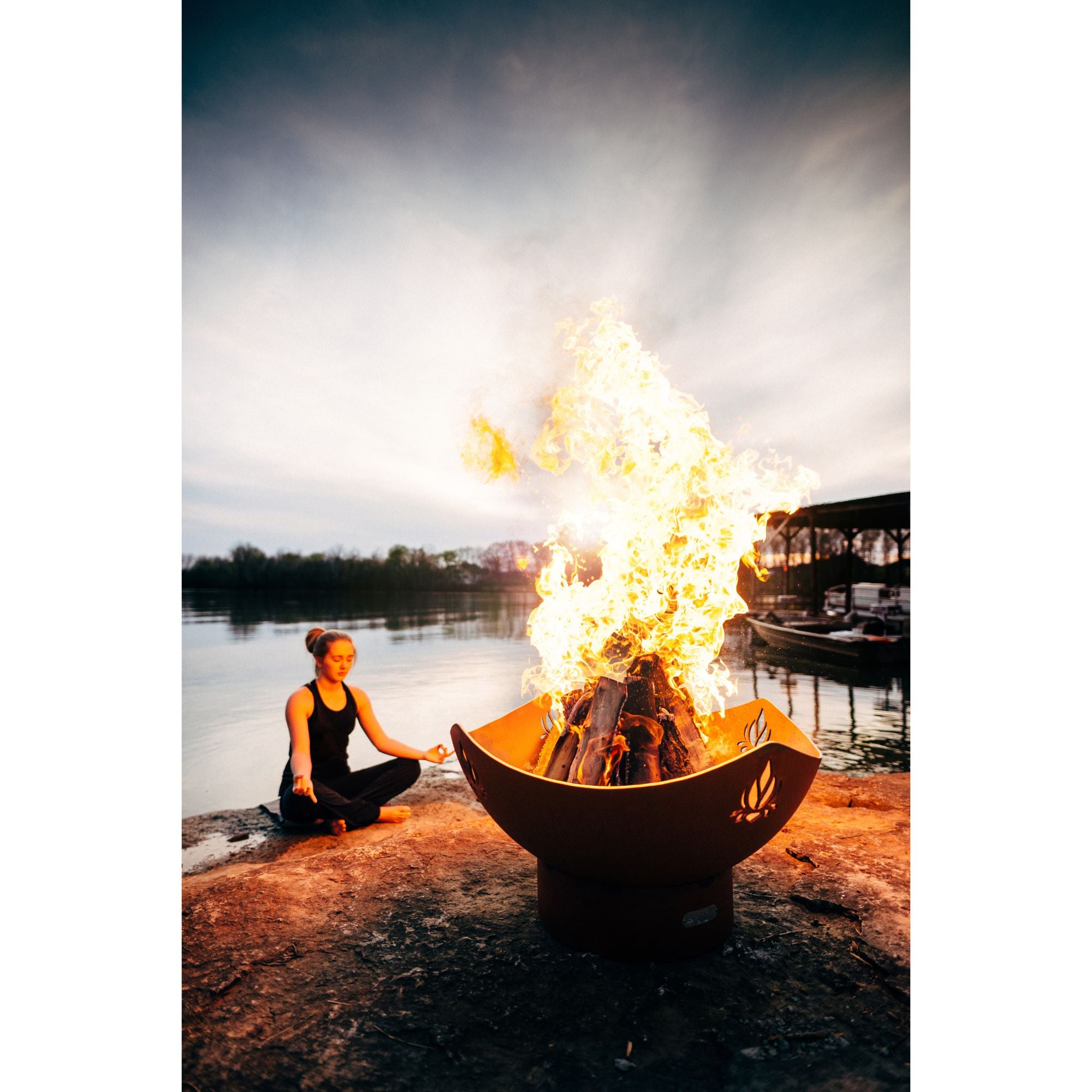 Person sitting by a fire pit by a lake with a boat in the background