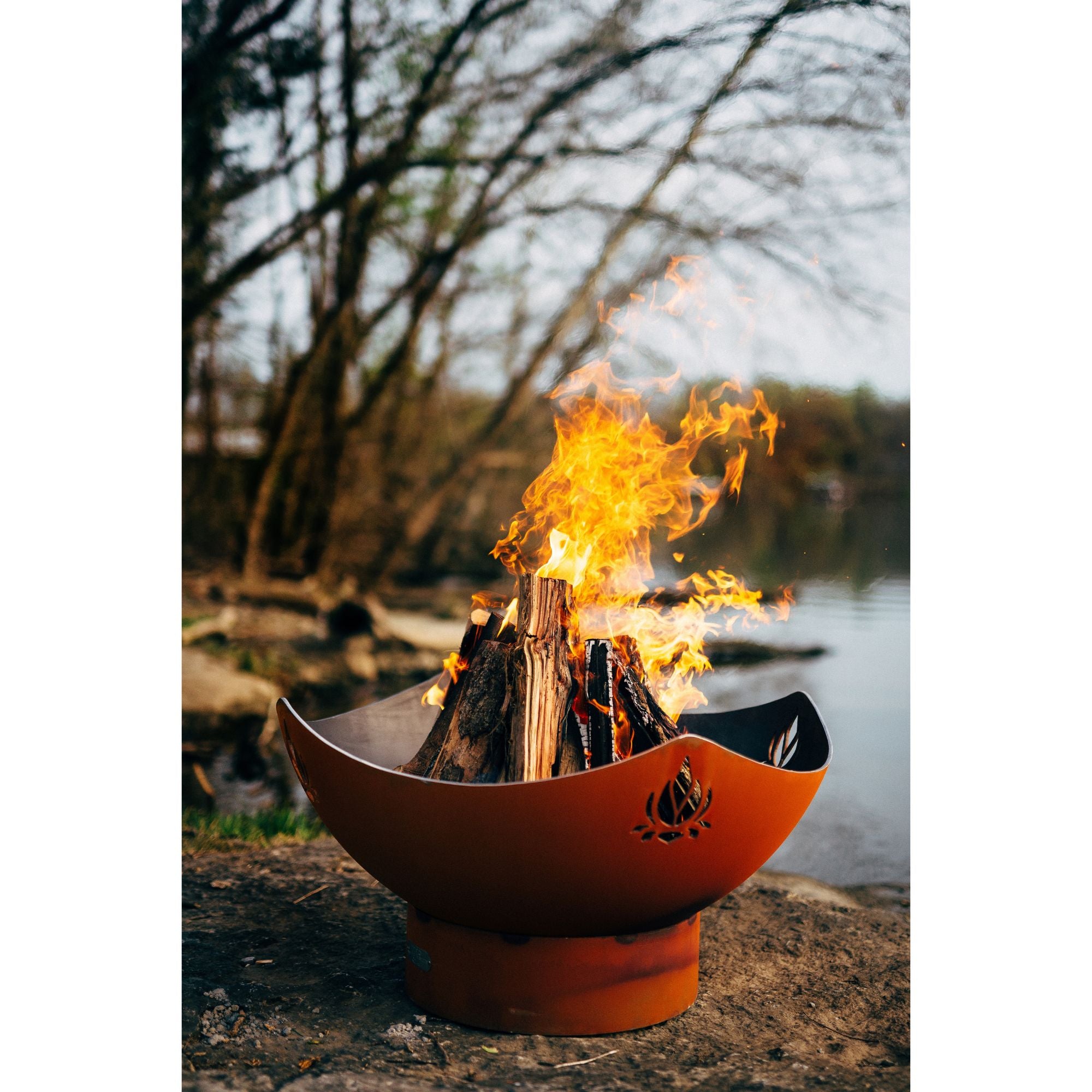 Orange fire pit with flames burning outdoors near a body of water.