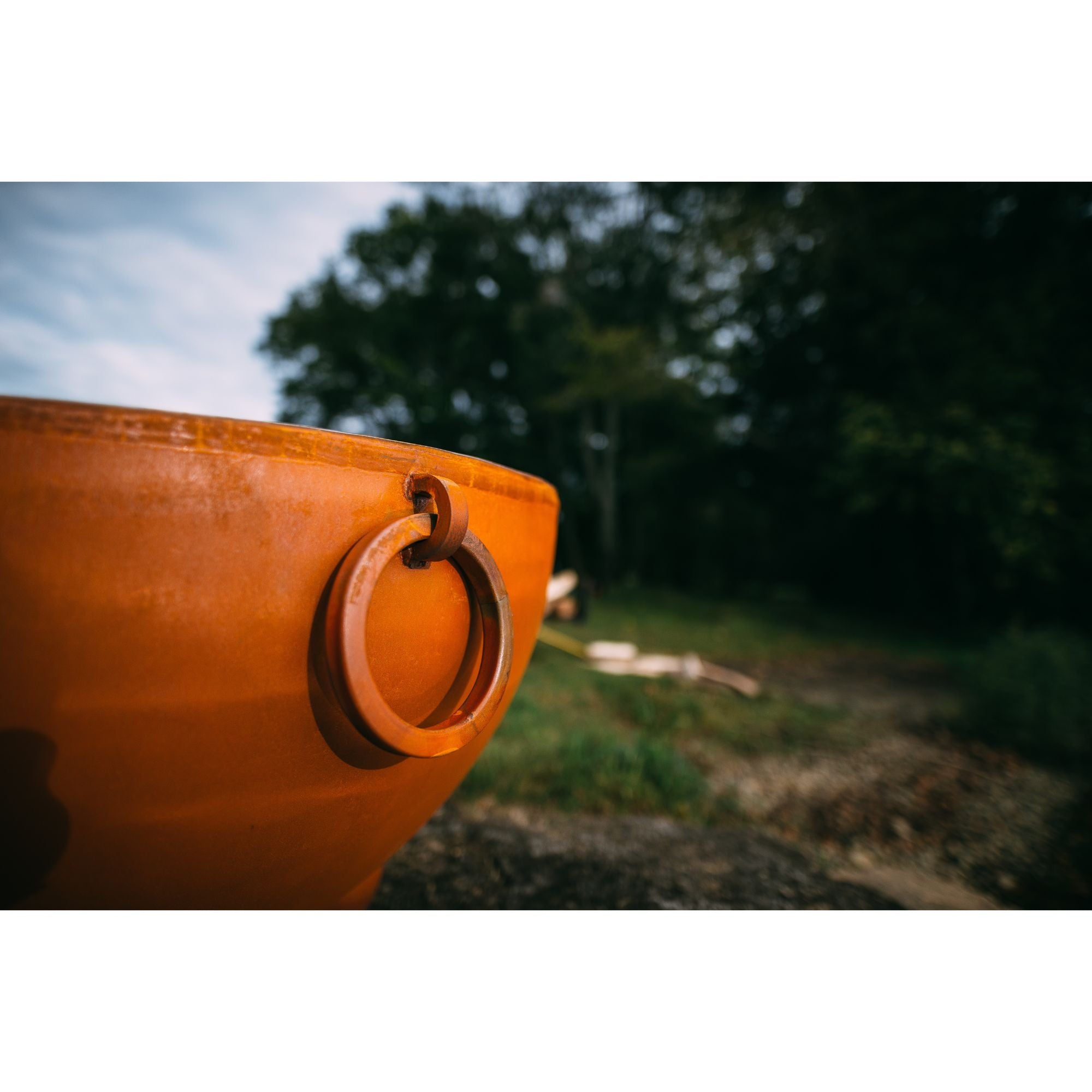 Close-up of an orange fire pit with a handle outdoors, blurred trees in the background
