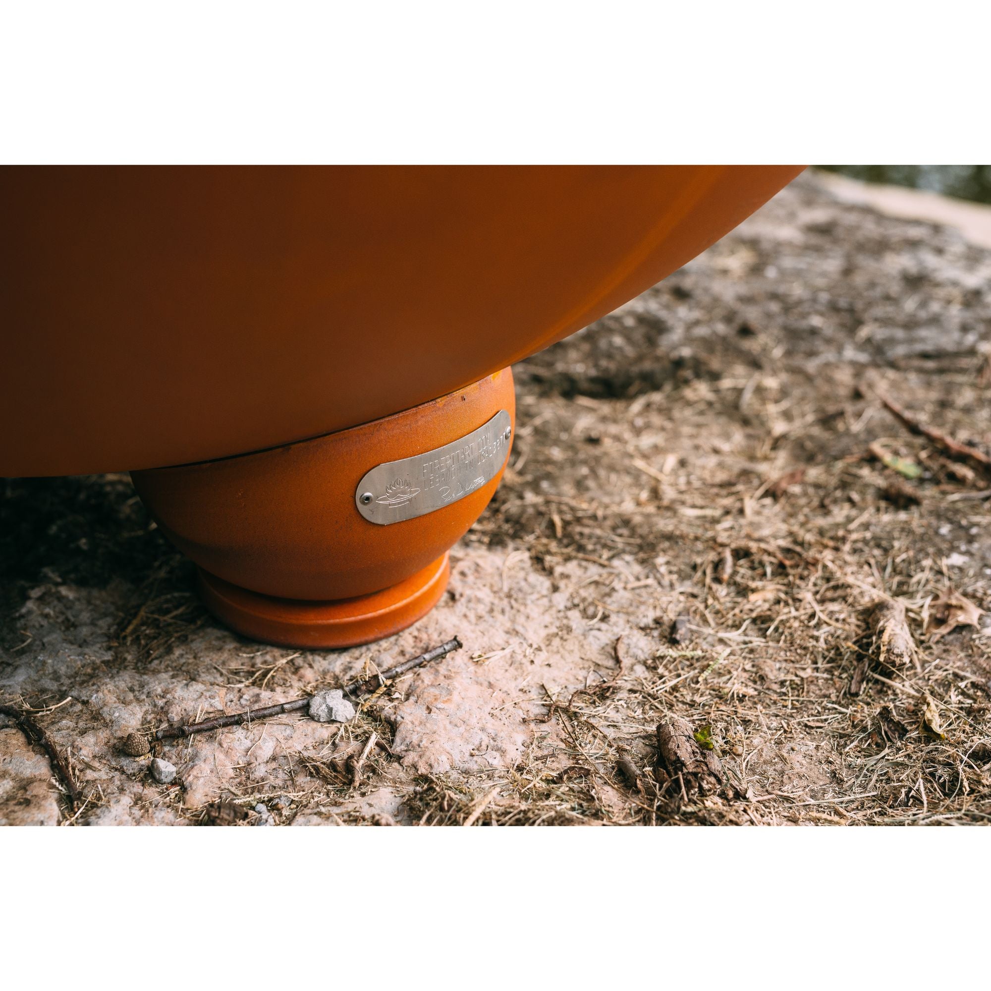 Close-up of a rust colored fire pit with a silver tag on a natural ground surface.