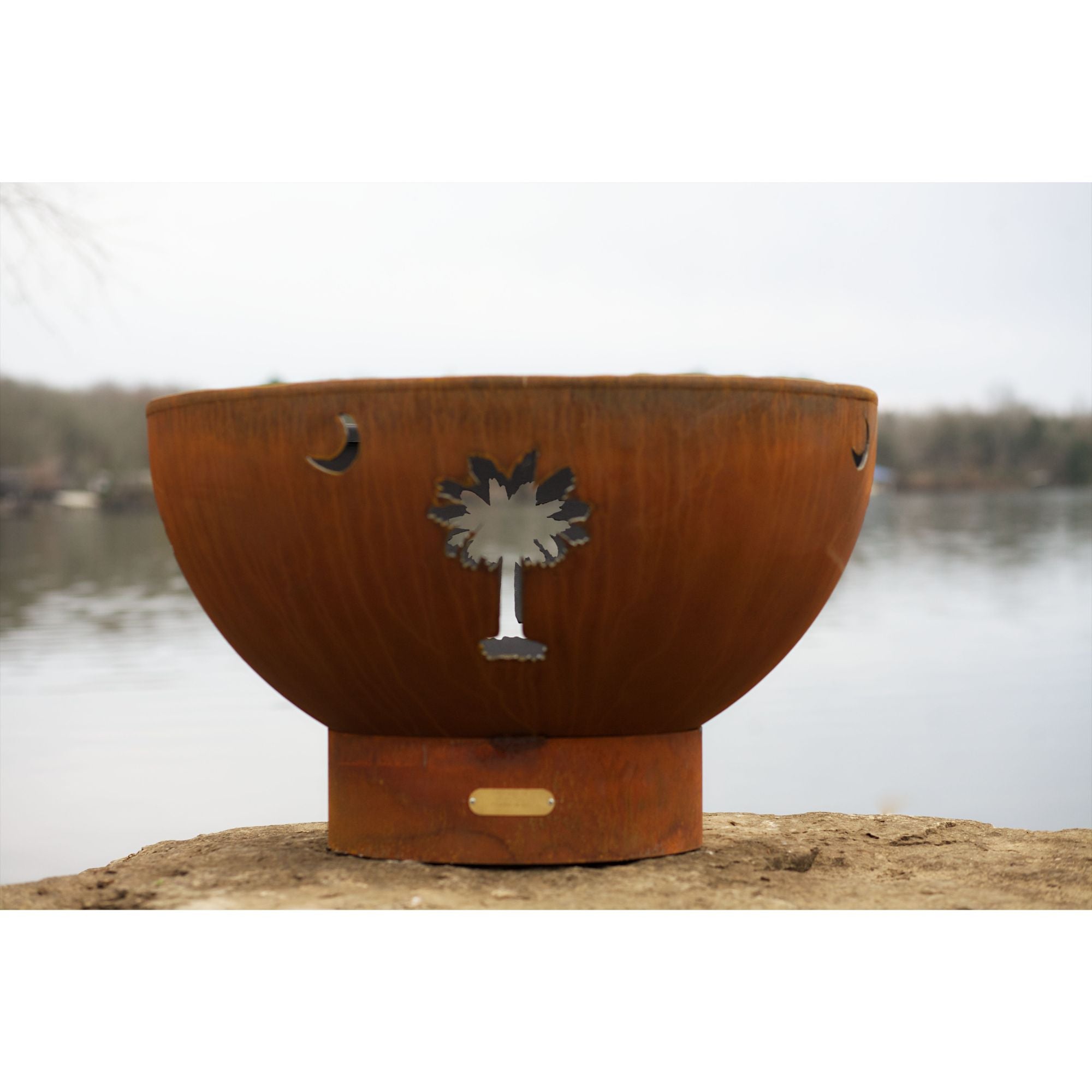 Rust-colored metal fire pit with a palm tree design on a wooden platform by a body of water.
