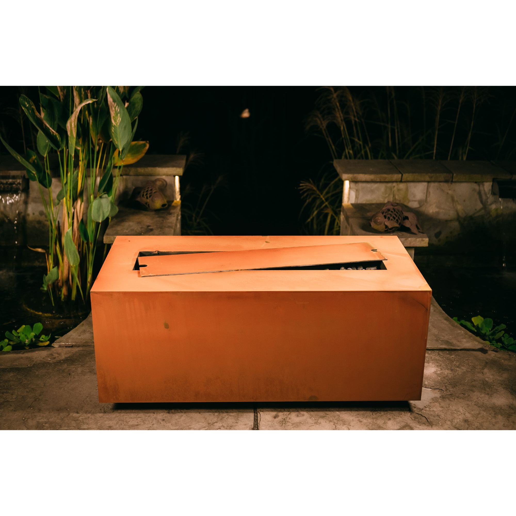 Rectangular fire pit in a garden setting with plants and a stone wall in the background.