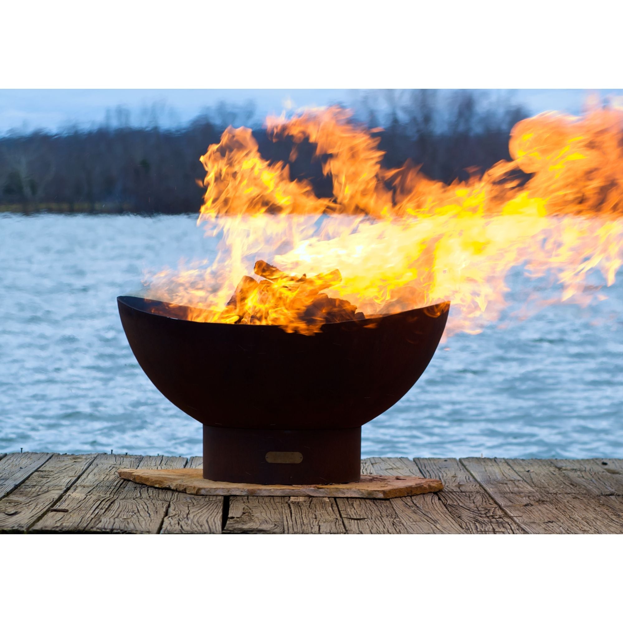 Fire pit with flames burning brightly on a wooden deck by a body of water.