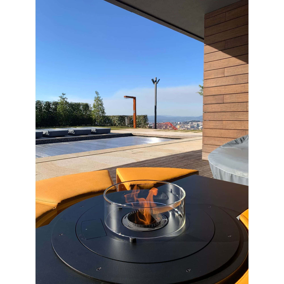 A modern outdoor fire pit table with a clear glass wind guard and visible flame, set on a patio next to orange seating under a covered area with a pool and sky in the background.