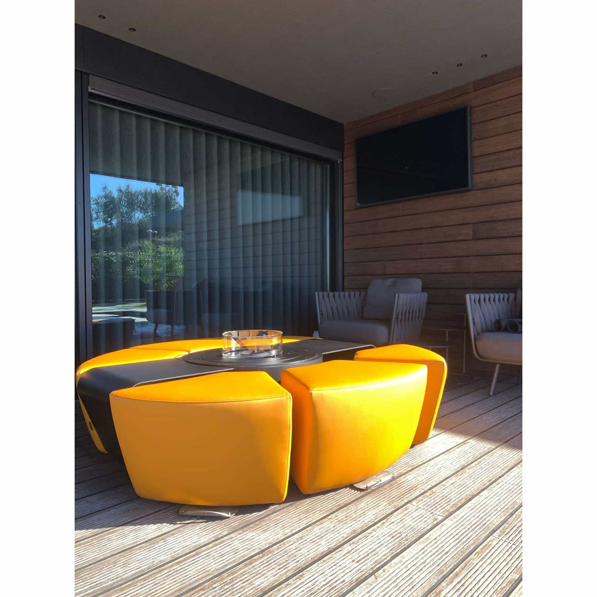 A modern outdoor patio seating area featuring a bright yellow modular curved sofa with a round black fire pit table in the center, set on a wooden deck with gray armchairs and a wood-paneled wall in the background.