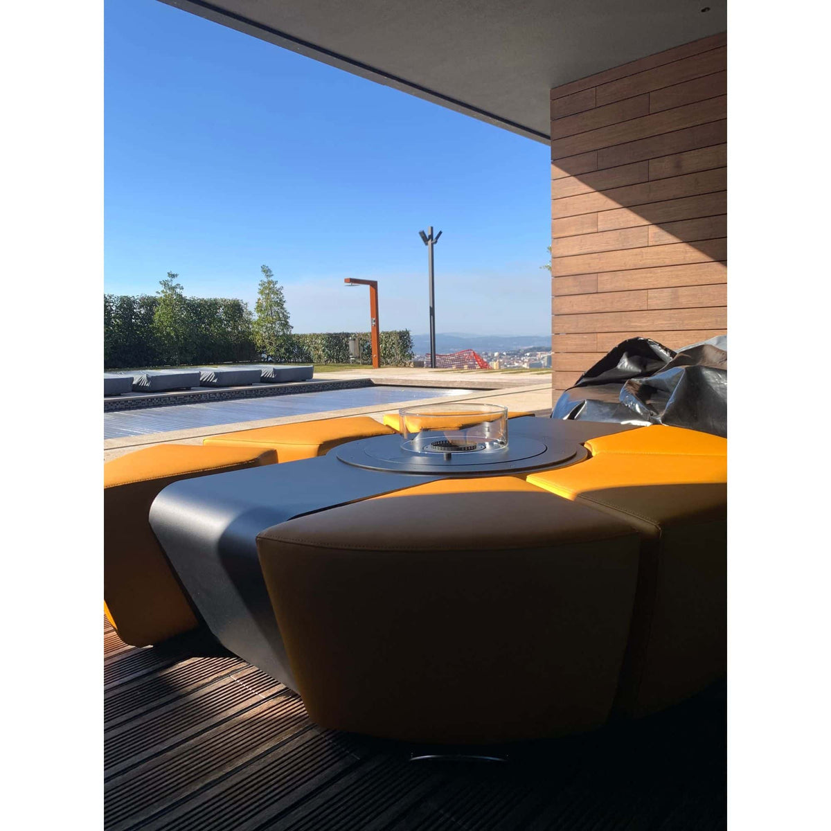 Modern outdoor seating area with curved orange upholstered seating around a fire pit table under a covered patio, with a terrace and trees visible in the background.