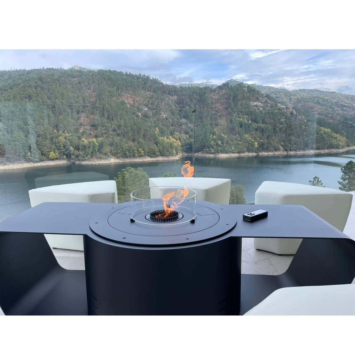 A modern outdoor patio fire pit table with a cylindrical black base and a glass wind guard, featuring a visible orange flame. The table is set on a patio with white lounge chairs, overlooking a scenic lake and forested hills.