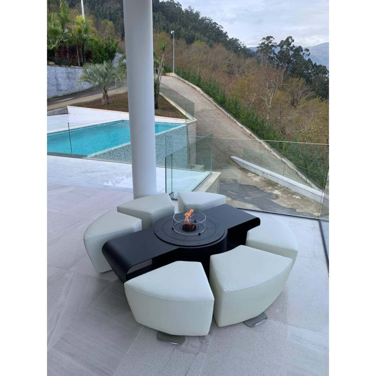 A modern outdoor patio furniture set featuring a black circular fire pit table with a central flame, surrounded by six curved white upholstered seat pods, set on a tiled patio with a pool and mountainous landscape in the background.