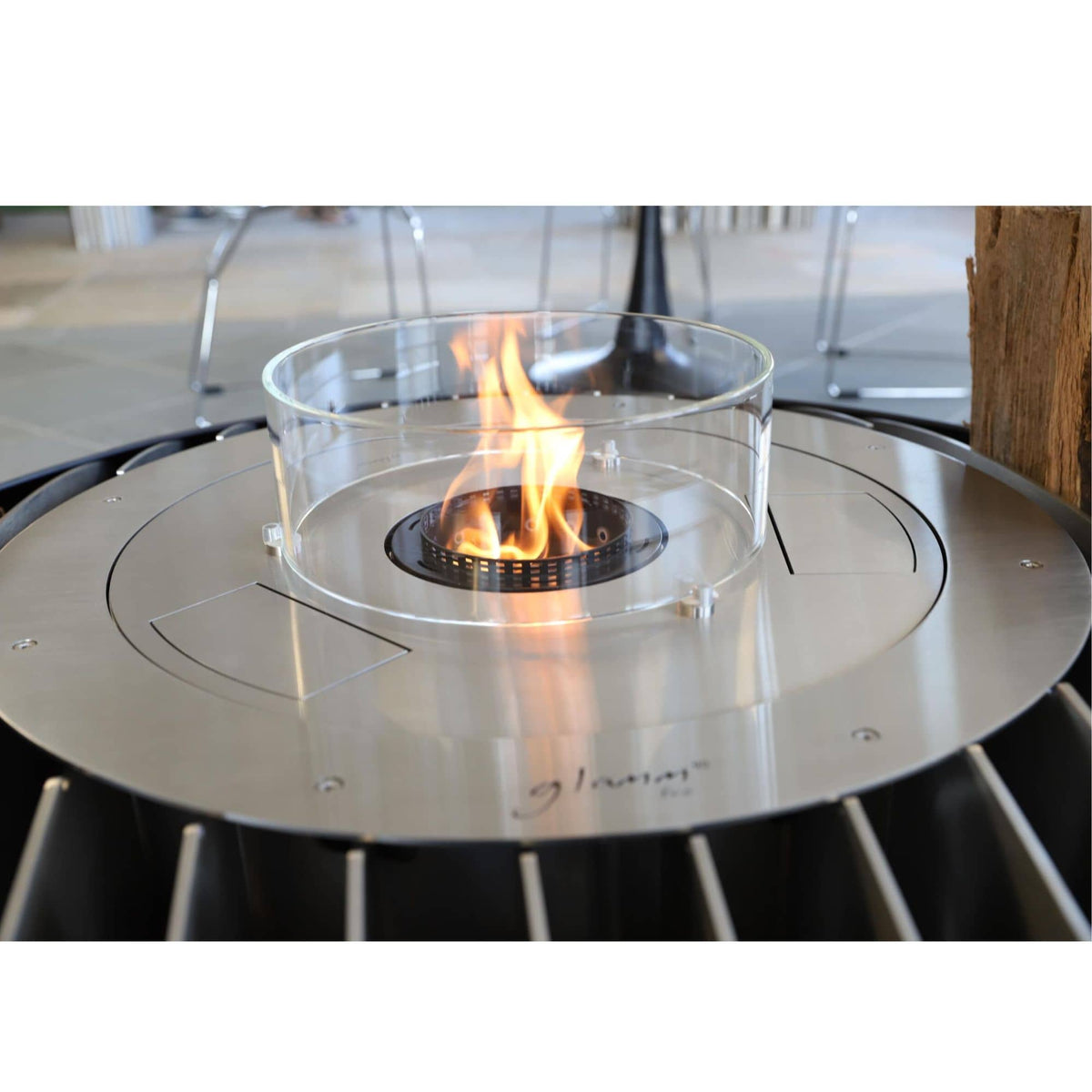 A close-up of a circular stainless steel outdoor fire pit with a clear glass wind guard and an active orange flame. The brand name 'glammfire' is visible in script on the front metal plate.