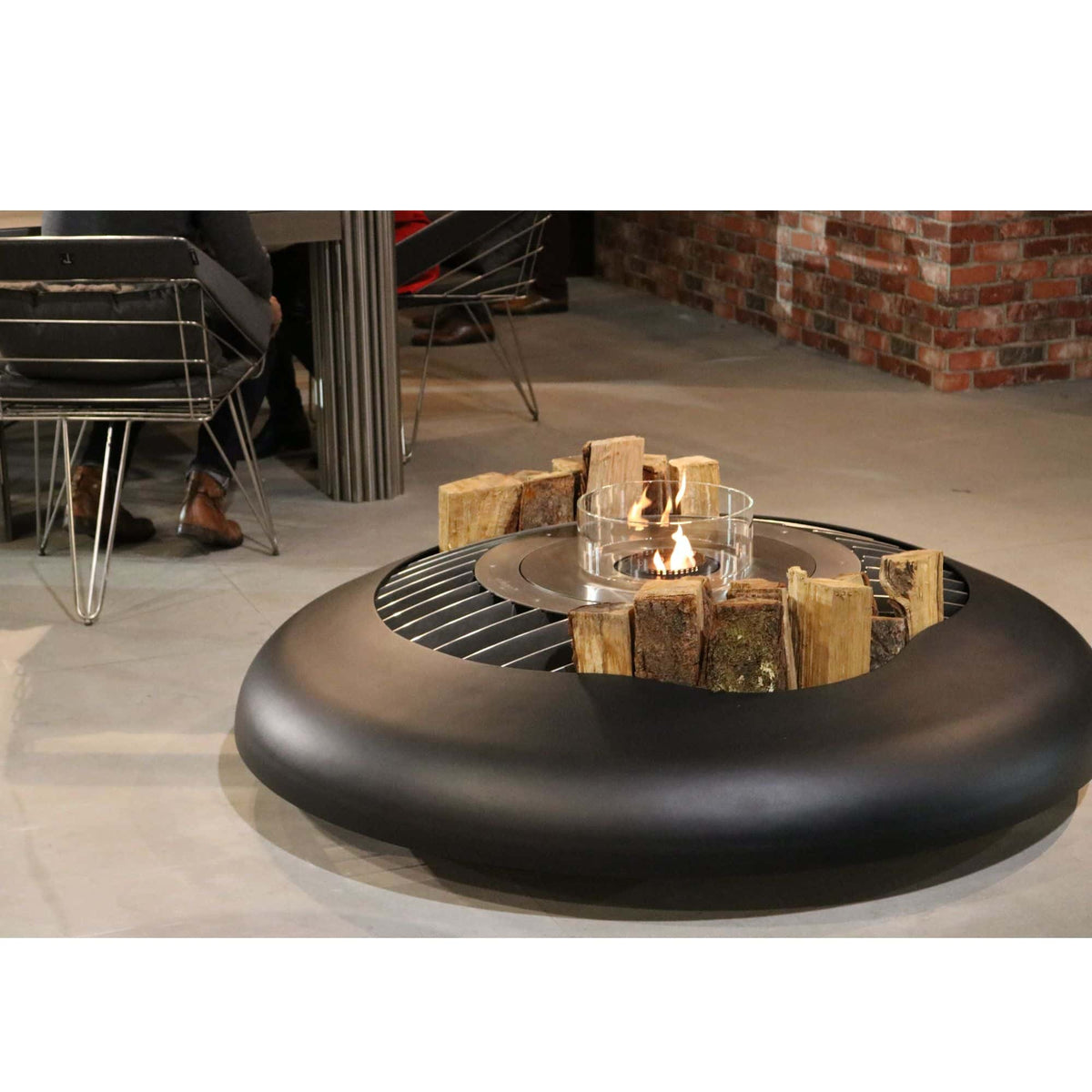 A round black outdoor fire pit table with a glass wind guard and decorative wood logs, shown in a modern patio setting with a brick wall and chairs in the background.