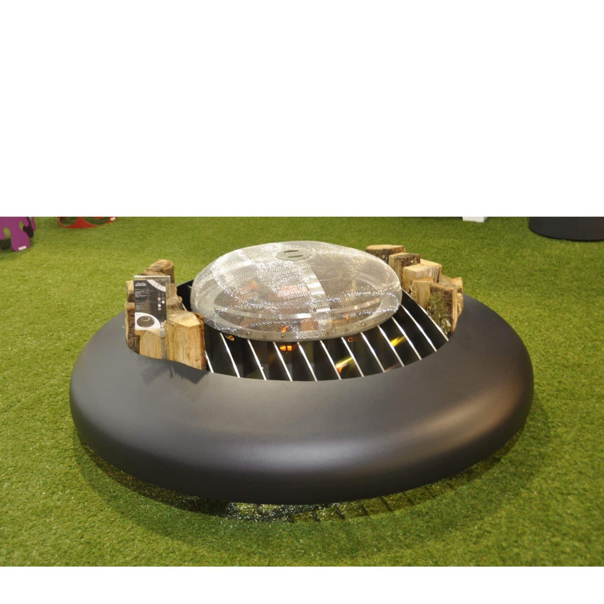 A round outdoor fire pit table with a black circular base, metal grates, small wood logs, and a clear domed lid, shown on artificial green turf.