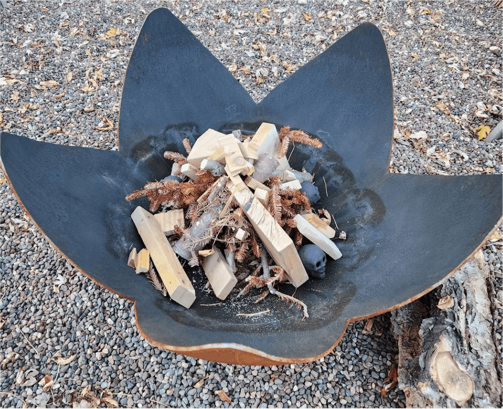 Black metal fire pit shaped like a flower with wood and twigs inside on a gravel surface.