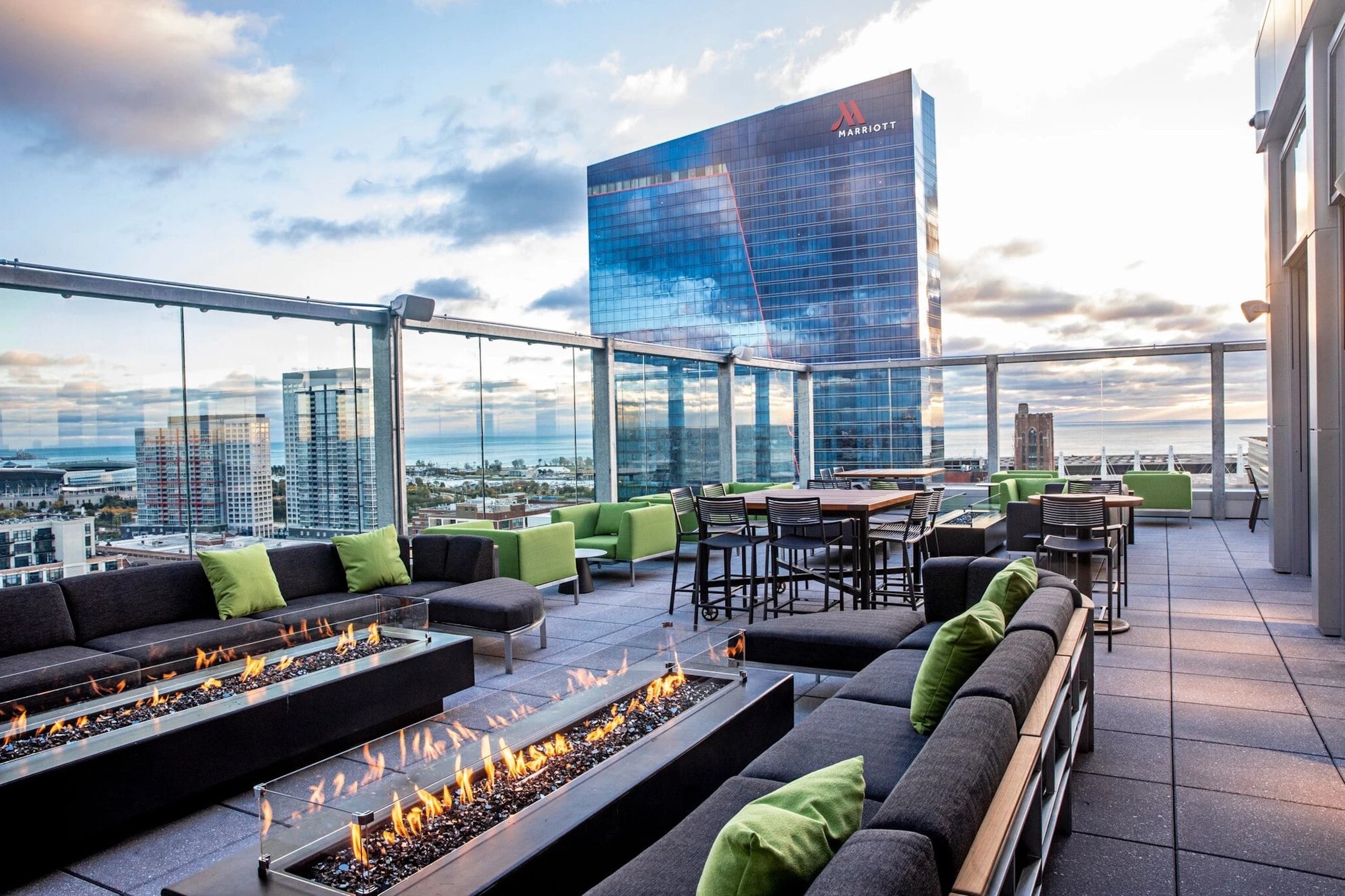 Outdoor rooftop lounge area with fire pit, tables, and chairs with a cityscape background.