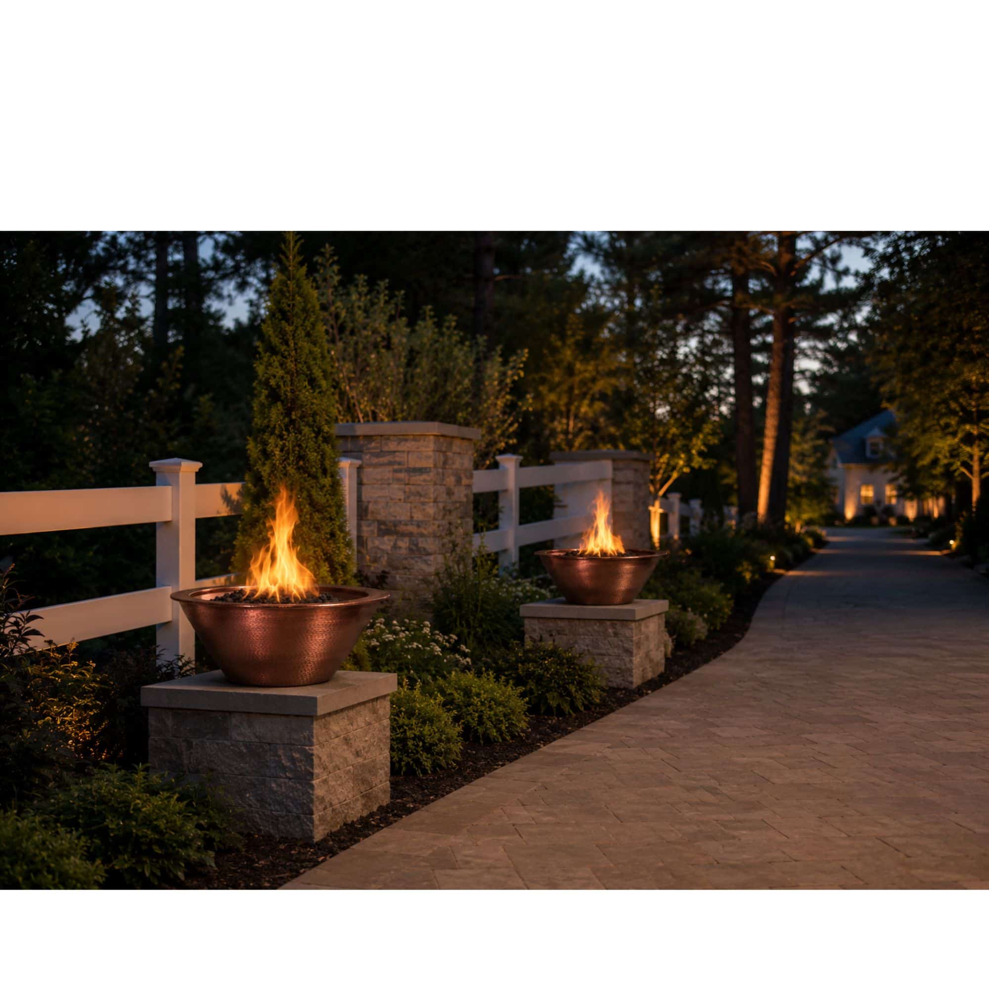 Two copper fire pits with active flames, set on stone pedestals along a paved pathway next to a white fence and stone pillar in a landscaped outdoor setting at dusk.