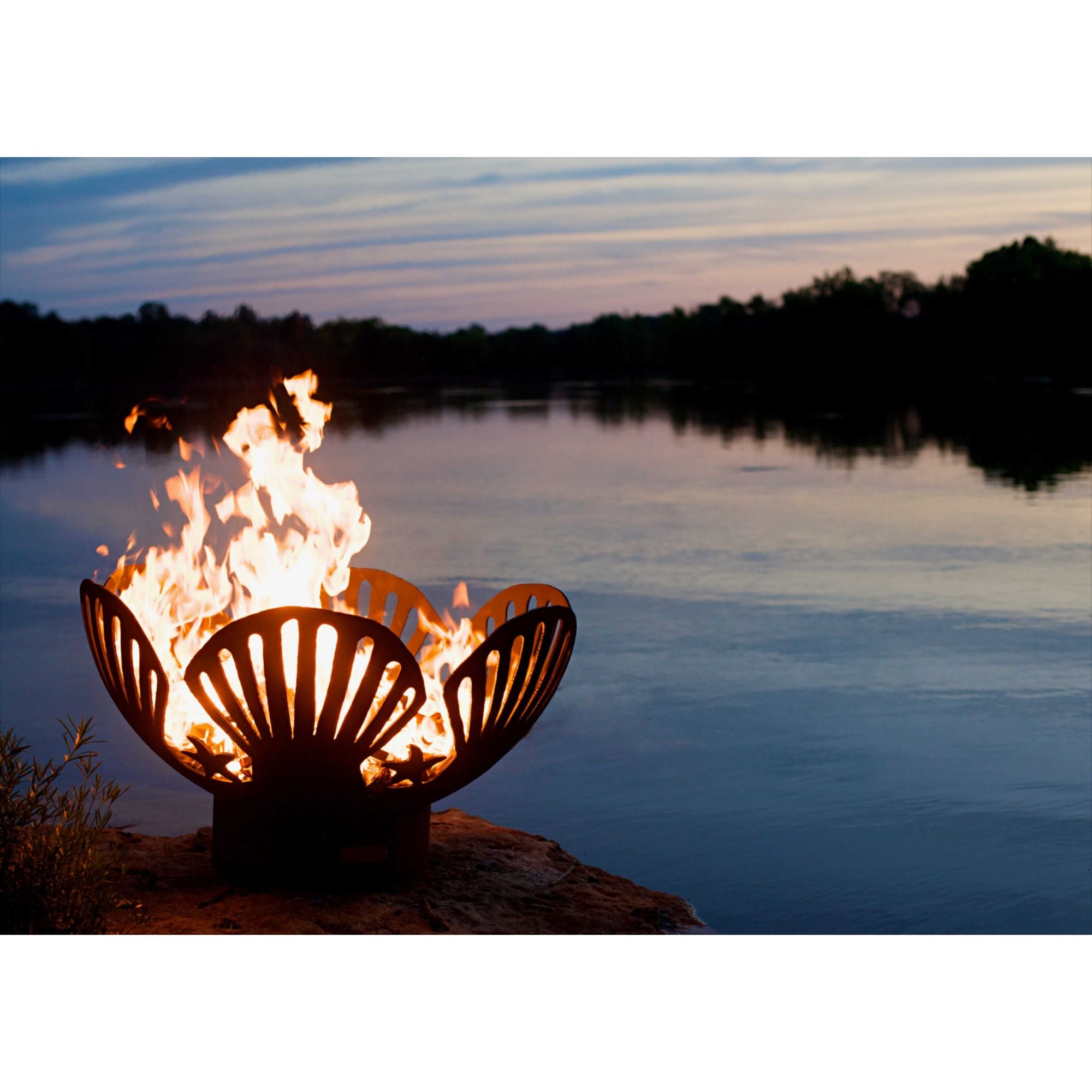 Fire pit with flames glowing brightly against a backdrop of a lake at dusk.