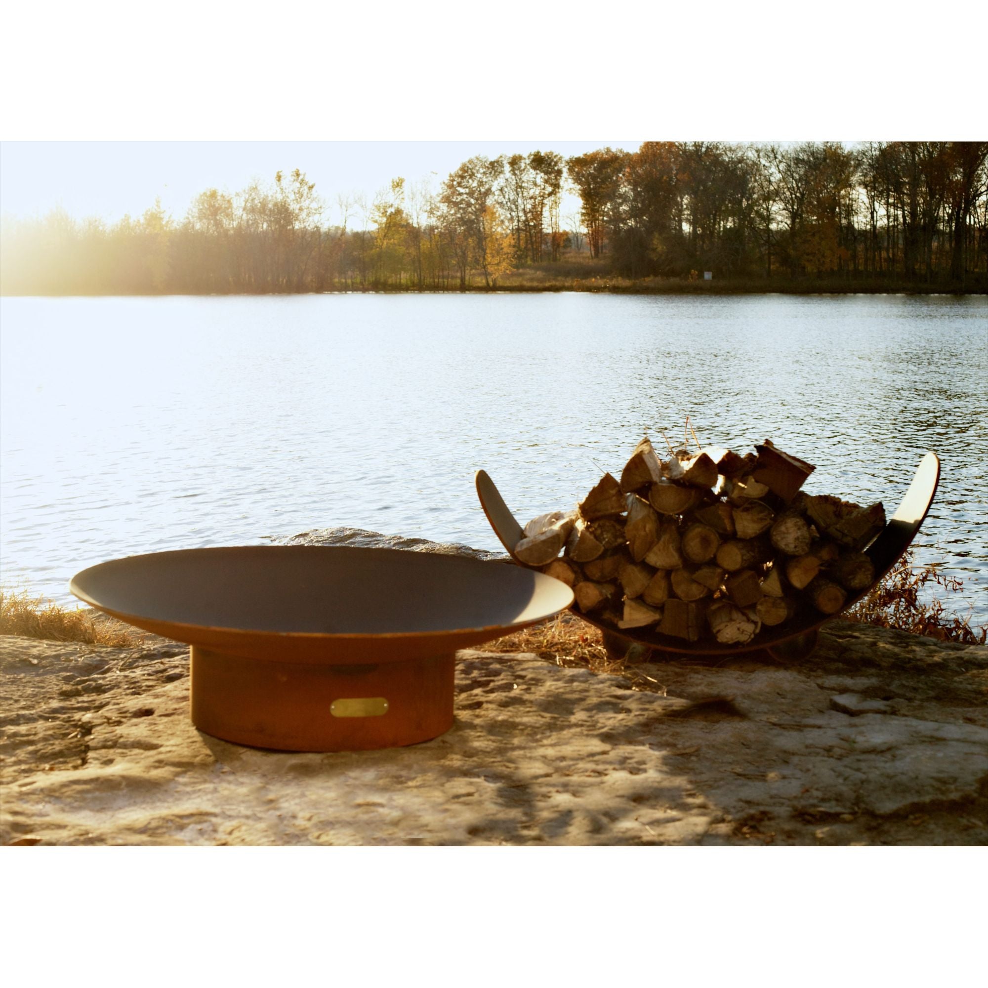 Fire pit and stacked logs by a lake with trees in the background