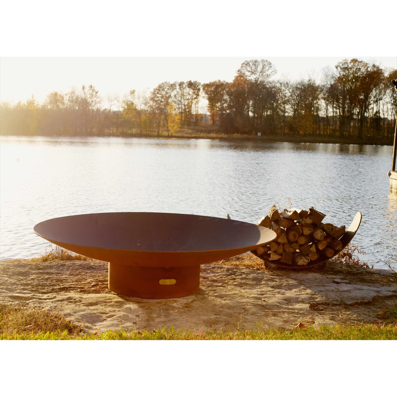 Rust-colored fire pit by a lake with stacked logs nearby