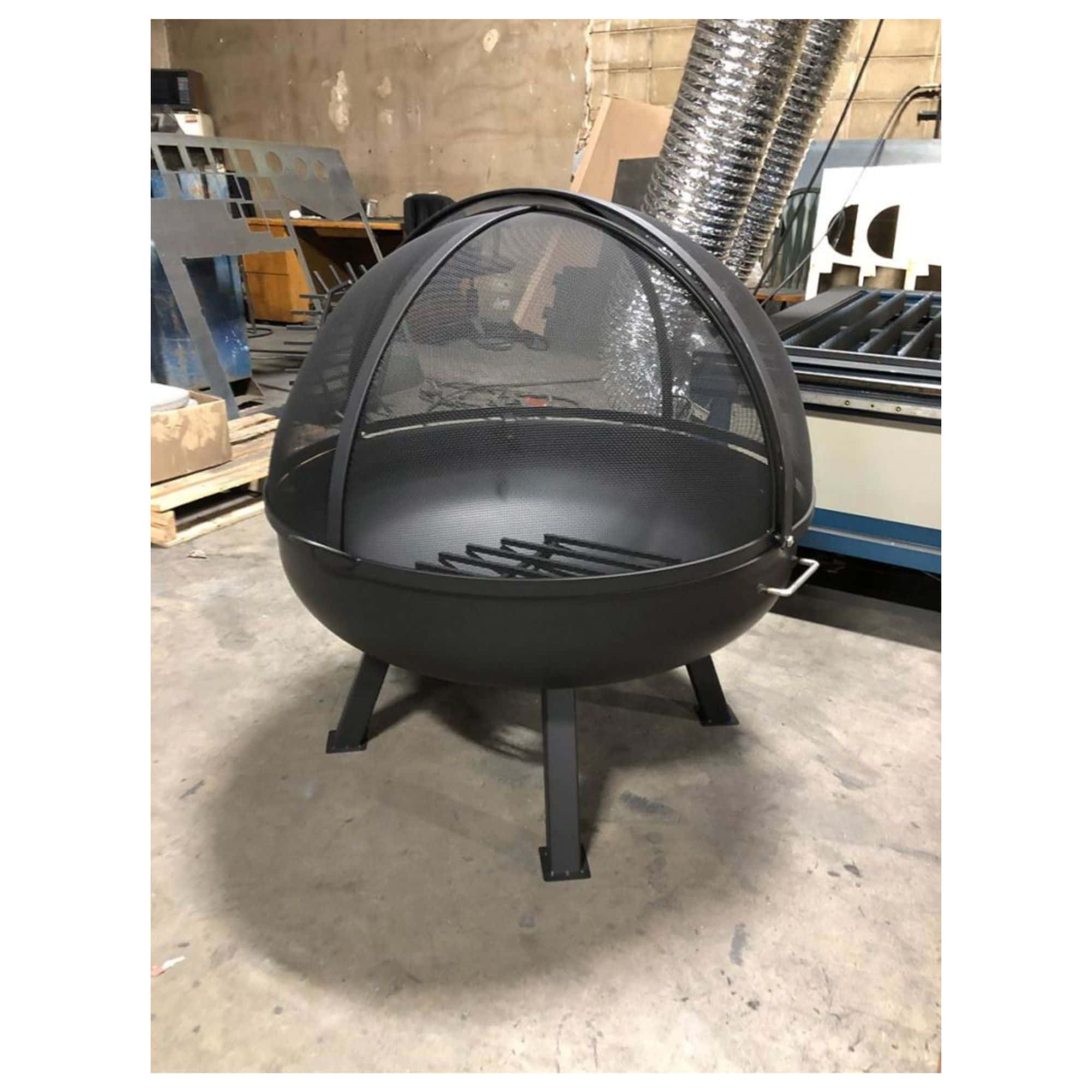 A black metal fire pit with a dome-shaped mesh screen and three legs, shown in a workshop or warehouse setting.