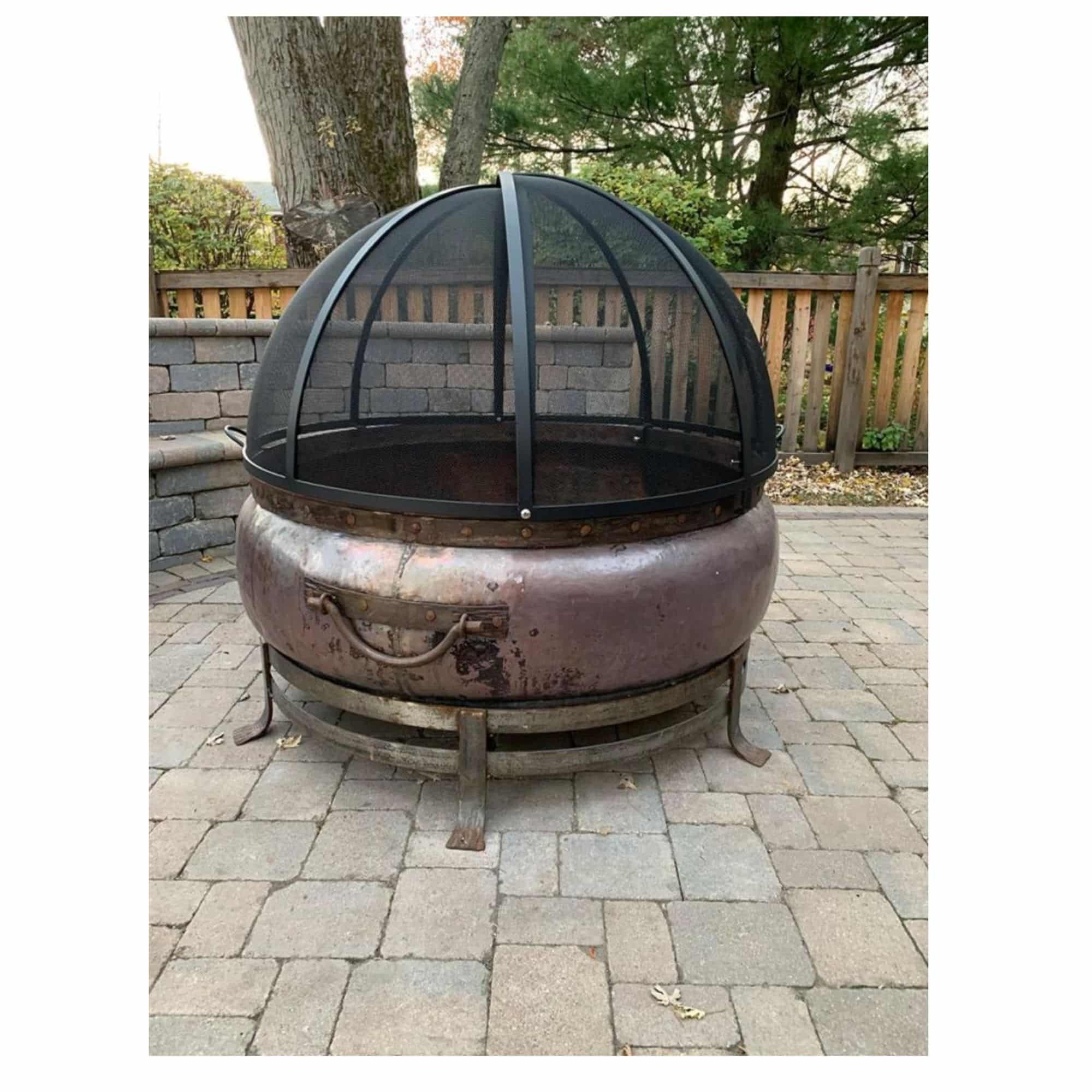 A round metal fire pit with a domed black mesh screen, set on a stone patio in an outdoor backyard environment.
