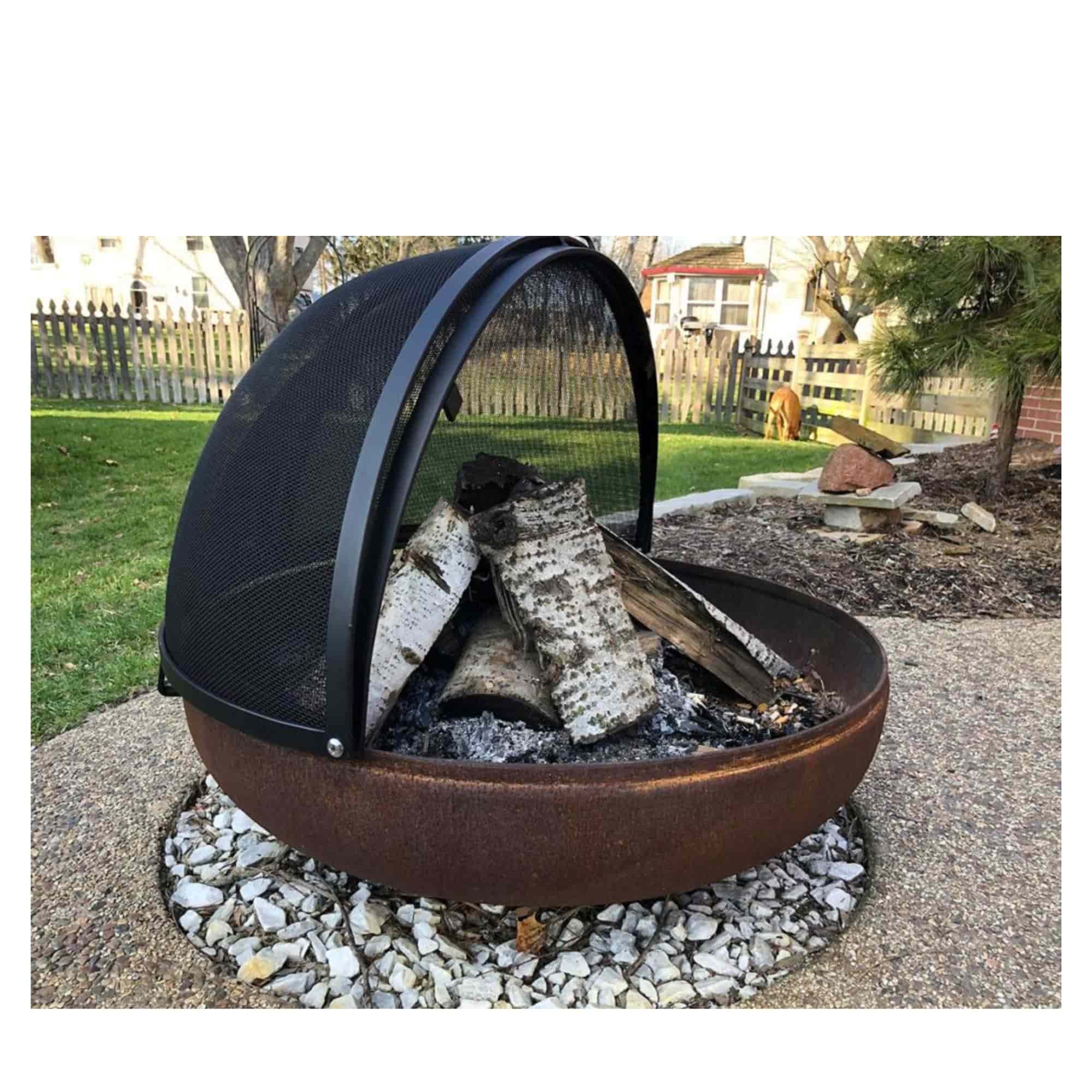 A round outdoor fire pit with a rusted brown metal bowl, black mesh dome cover, and stacked firewood, placed on white gravel in a backyard setting.