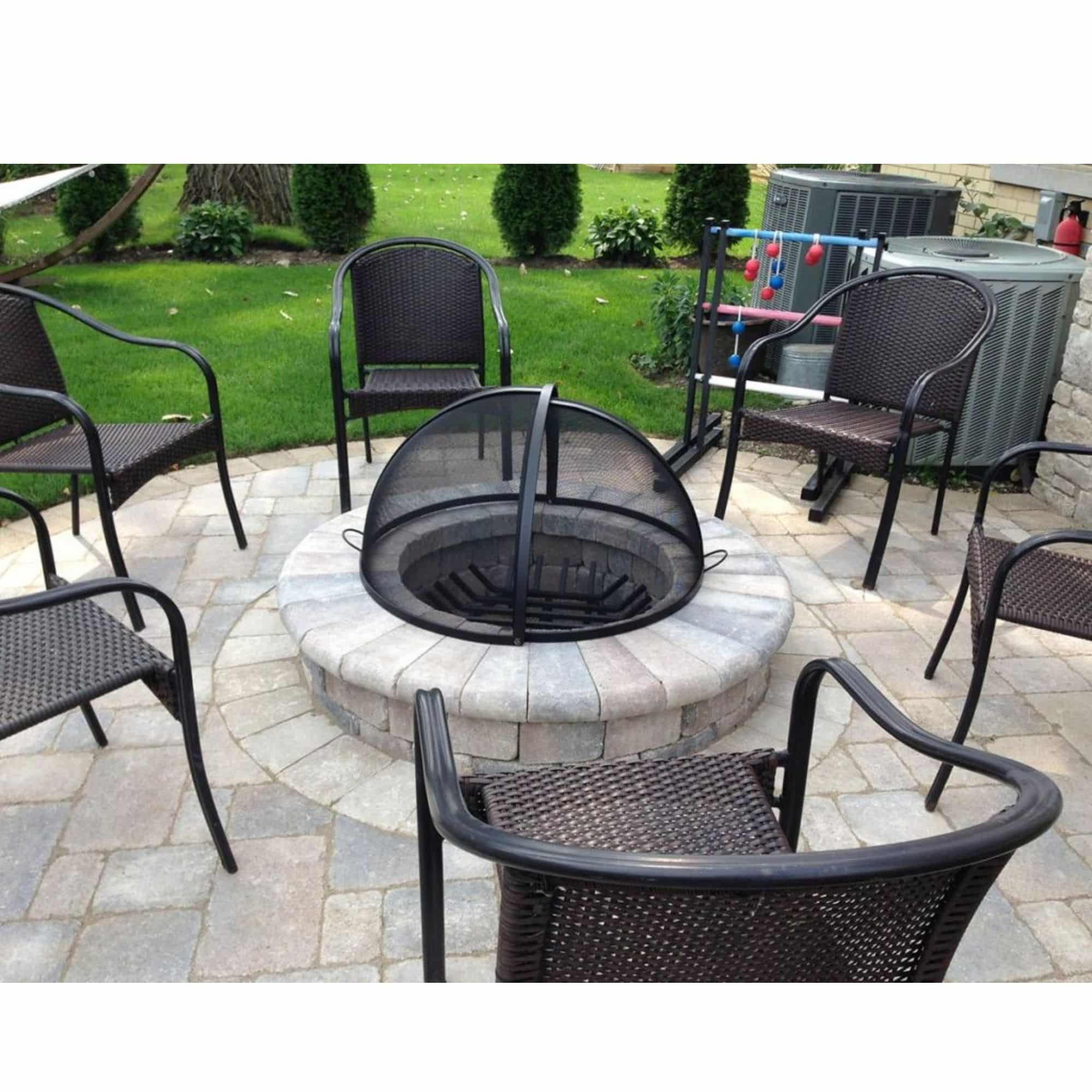 Outdoor patio fire pit with a black mesh screen cover, surrounded by dark woven patio chairs on a stone paver deck with a green lawn in the background.