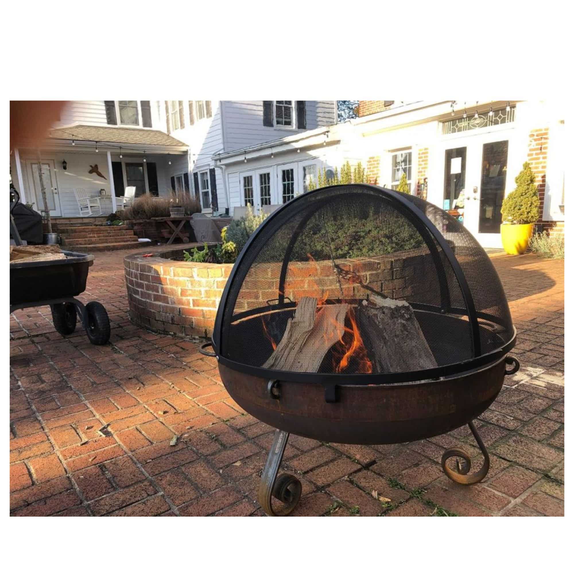 A round metal fire pit grill with a domed black mesh screen cover, shown outdoors on a brick patio with wood logs burning inside. Residential houses and a brick planter are visible in the background.