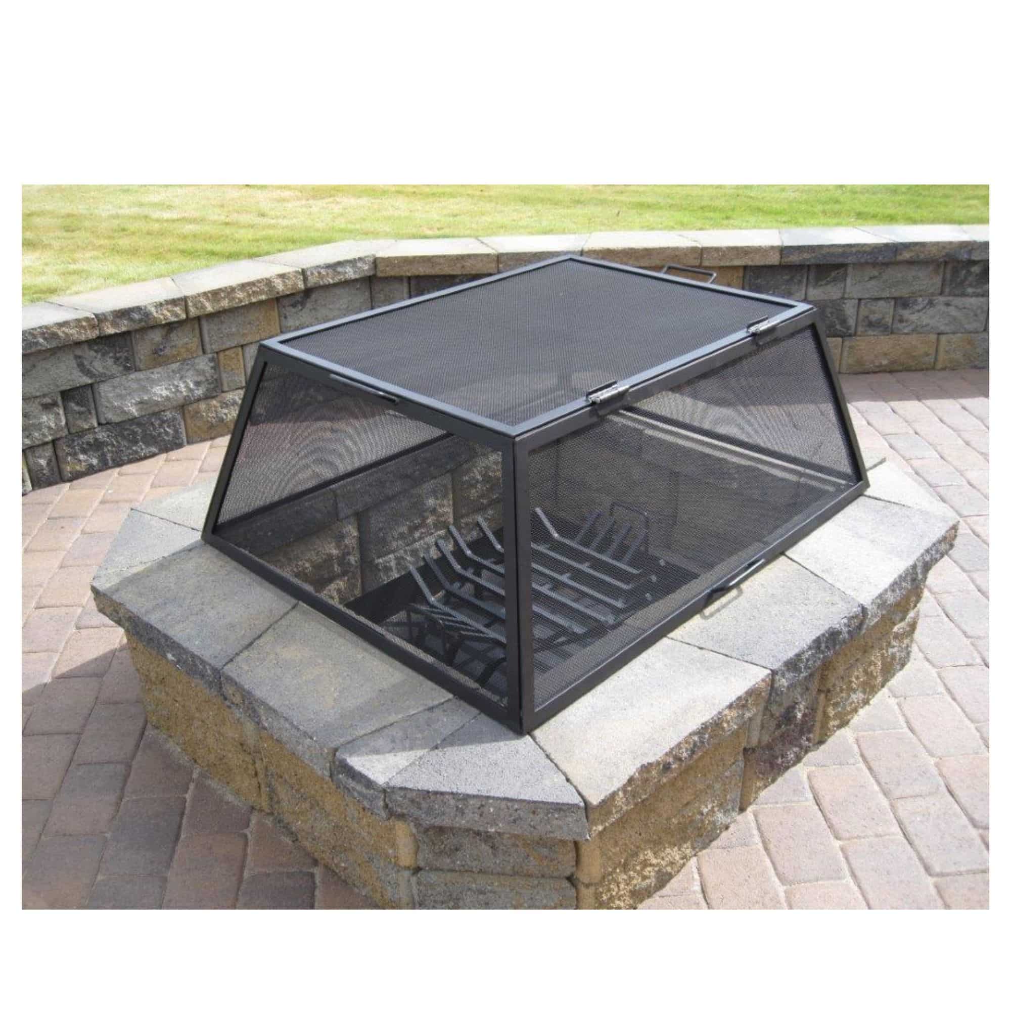 A black metal and mesh fire pit cover with a built-in fire grate, shown outdoors on a stone patio next to a brick wall and grass.