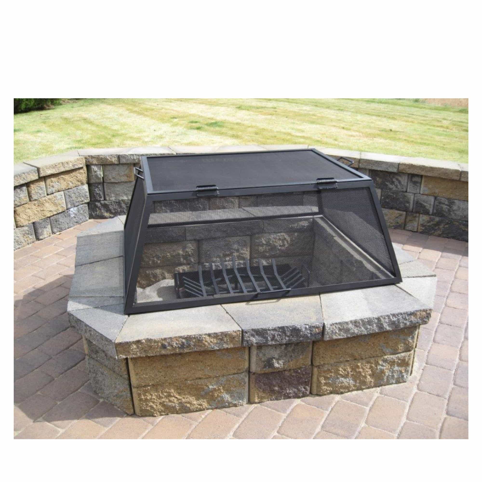 A black metal fire pit cover with a mesh screen lid and integrated fire grate, shown installed over a stone fire pit on a paved patio with a grassy lawn in the background.