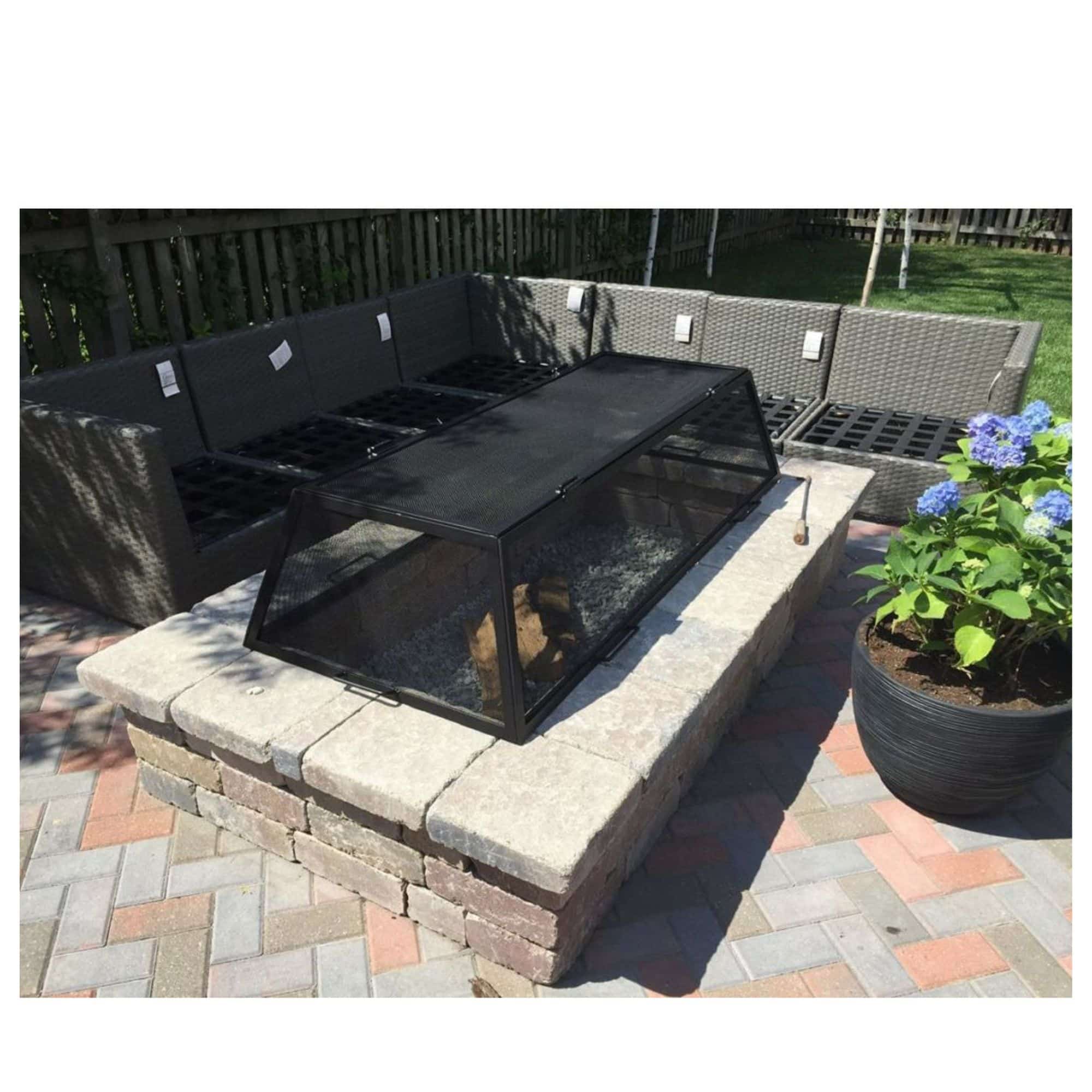 A black outdoor fire pit cover, shown on a brick patio next to a wicker patio sofa and a potted plant.