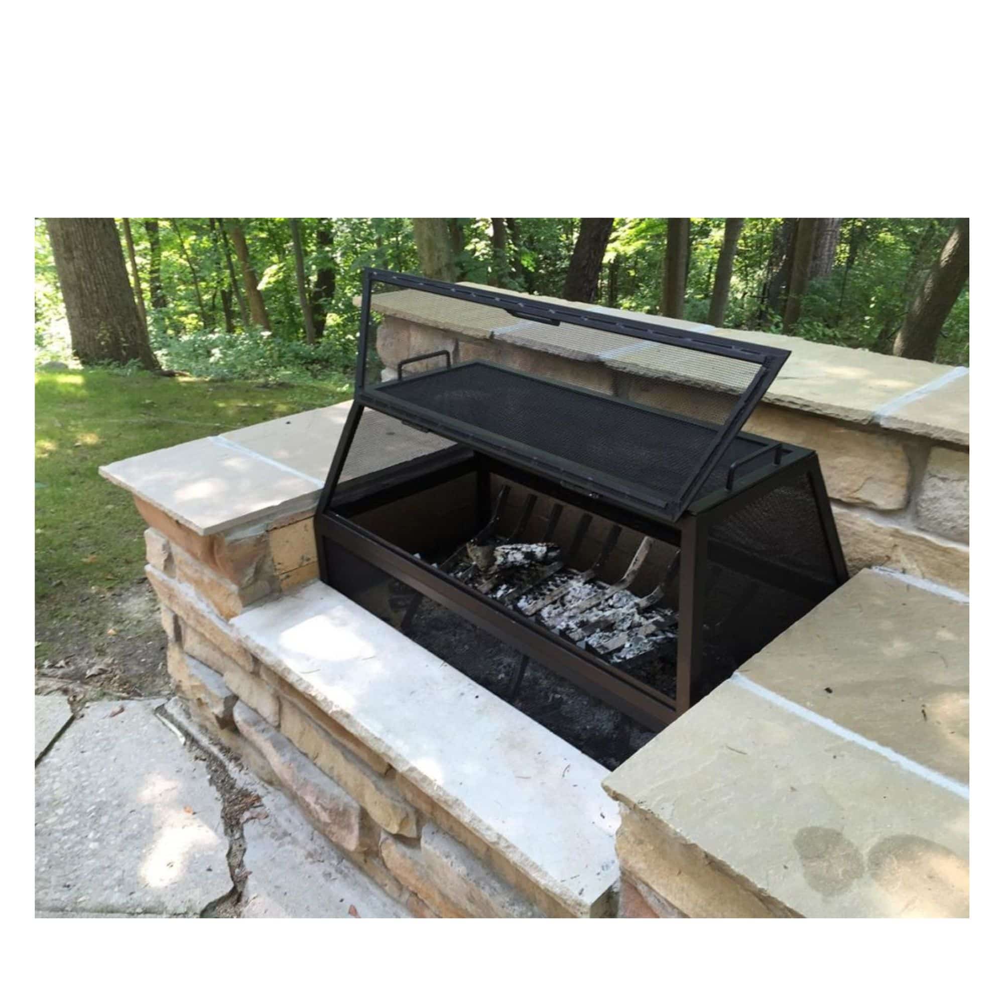 A black metal outdoor fire pit with a hinged lid, shown open with embers inside, set into a stone patio with a wooded background.