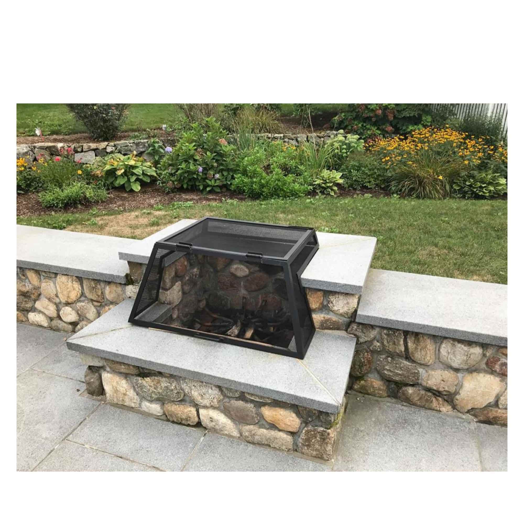 A black metal fire pit cover with a mesh screen and glass front panel, shown placed on a stone patio in an outdoor garden setting.