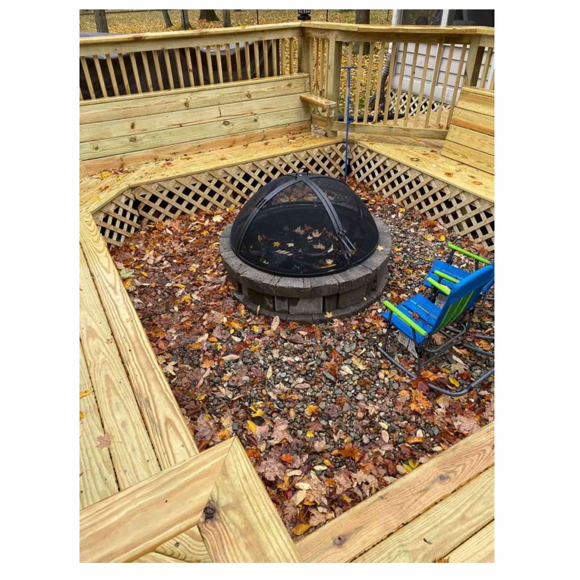Outdoor backyard scene showing a round fire pit with a black mesh cover, surrounded by a wooden deck and lattice railing, with fallen leaves on the ground and a blue chair on the right.