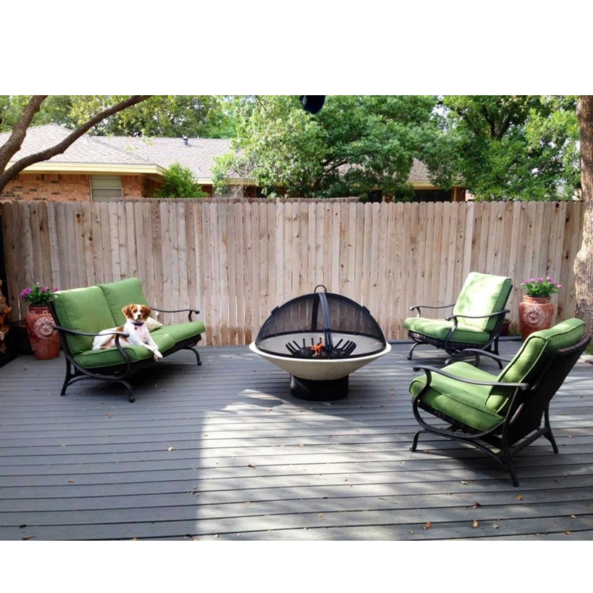 Outdoor patio furniture set on a gray deck with a wooden fence and trees in the background. The set includes two green cushioned lovese-style chairs with black metal frames, a matching green cushioned armchair, and a round light brown fire pit bowl with a black fire pit screen in the center.