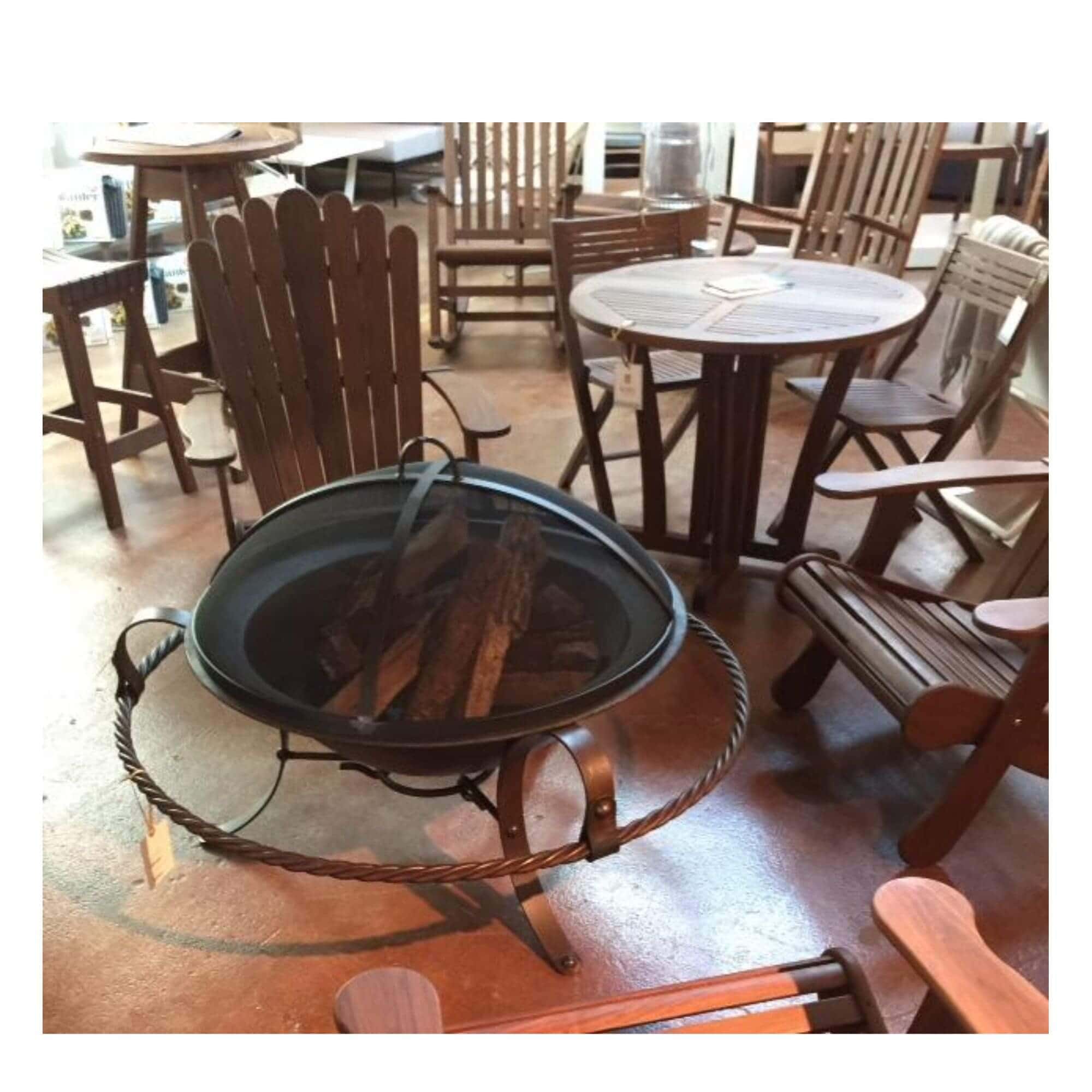 A dark metal fire pit table with a black fire pit screen with a rope-wrapped base and wood logs inside, photographed in a furniture showroom with other wooden chairs and tables around it.