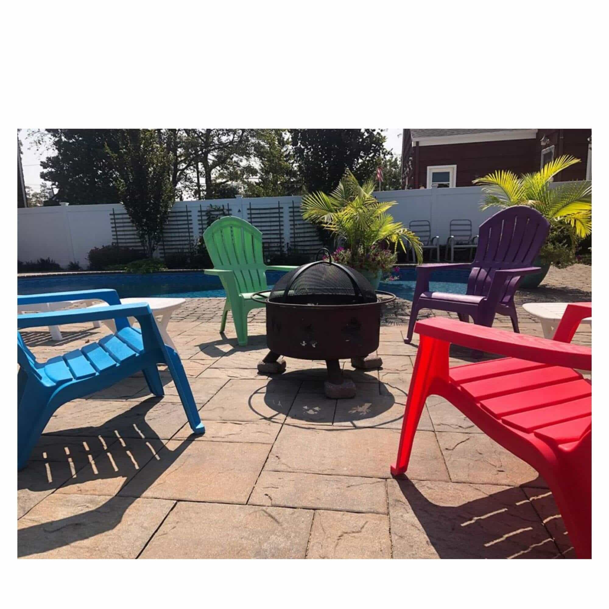 Four colorful outdoor Adirondack chairs in blue, green, purple, and red arranged around a black fire pit with a black fire pit screen on a stone patio next to a swimming pool.