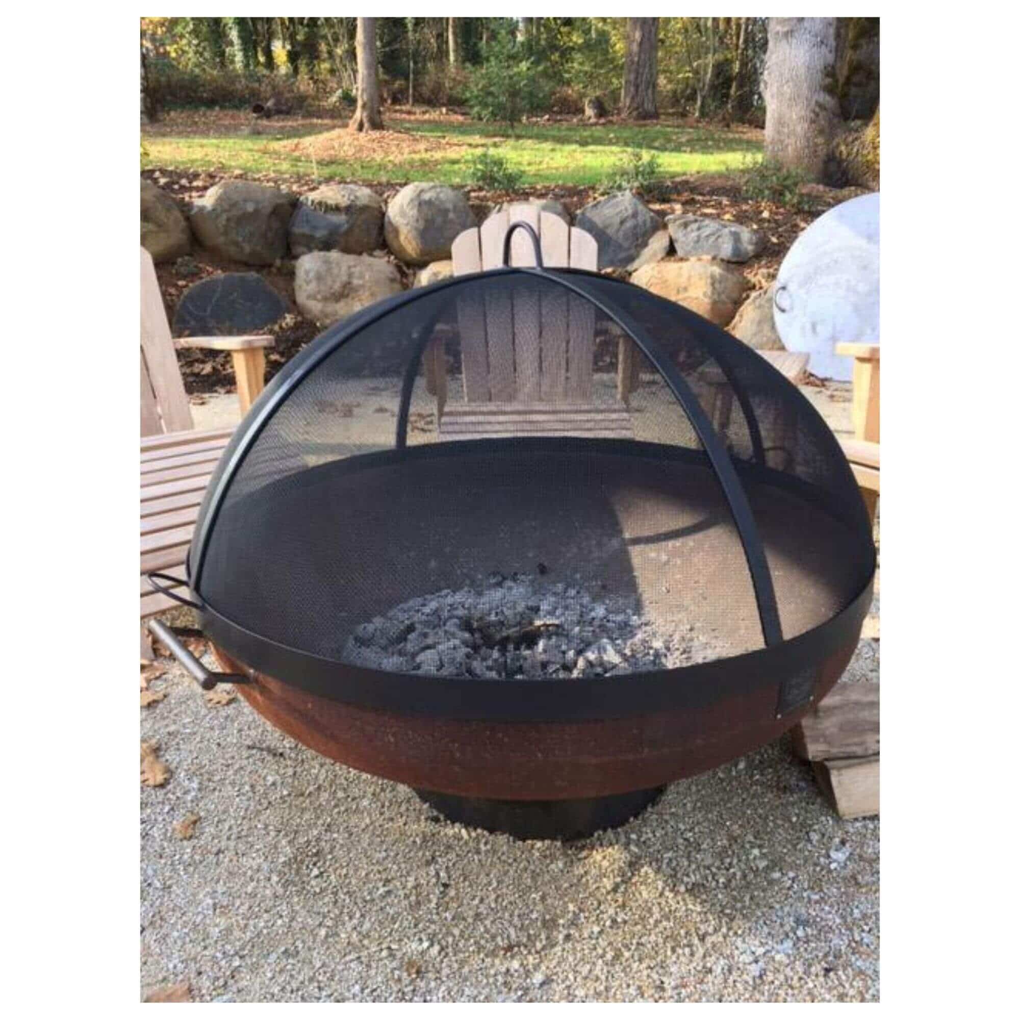 A round metal fire pit with a black mesh dome screen, shown outdoors on a gravel surface with patio chairs and trees in the background.