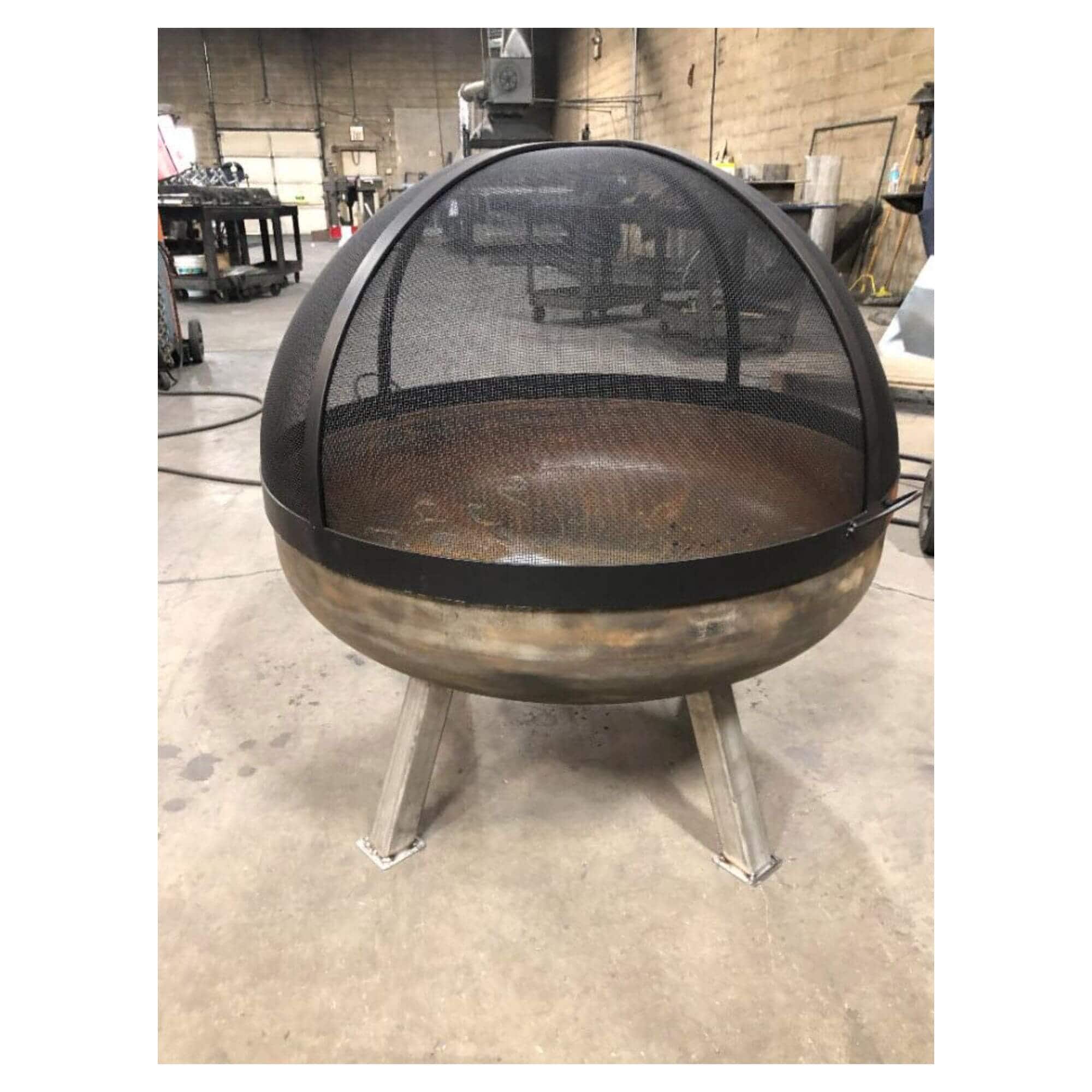 A round metal fire pit with a black mesh dome cover and three legs, photographed in an industrial workshop setting.