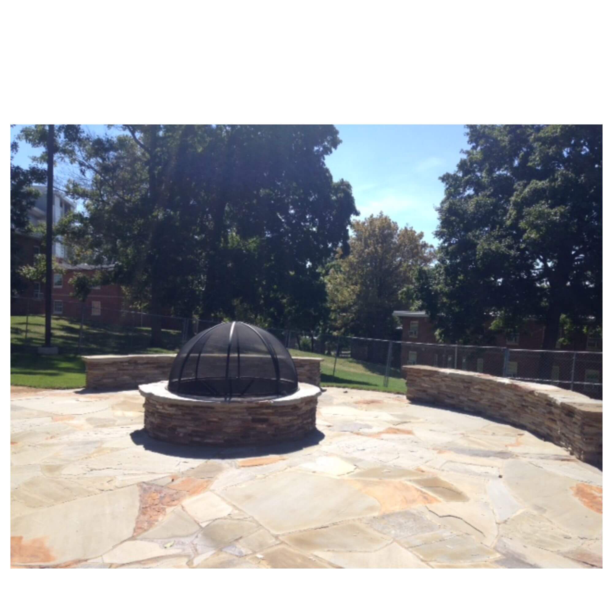 Outdoor stone patio with a round fire pit featuring a black dome cover, surrounded by low stone walls and trees in the background.