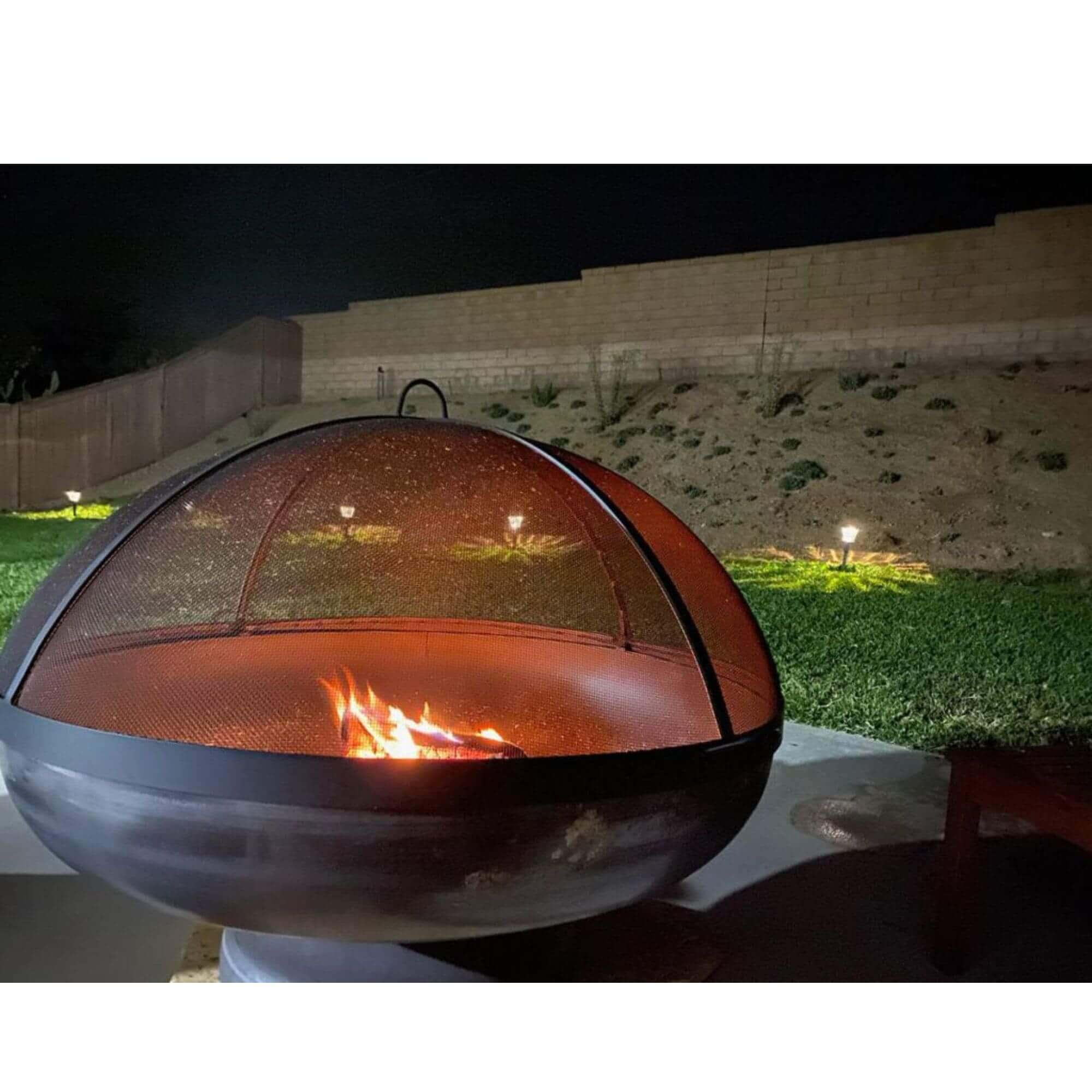 A spherical metal fire pit with a domed mesh cover and visible flames, placed outdoors on a patio at night with grass, a wall, and landscape lights in the background.